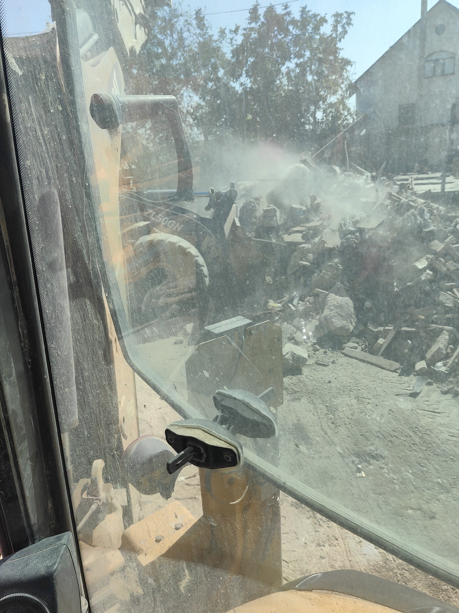 The image shows a construction site in Shymkent, Kazakhstan, viewed from inside the cab of a yellow excavator. The foreground is dominated by the dusty windshield of the excavator, with a rearview mirror and a control lever visible. Through the windshield, a chaotic pile of rubble, including bricks, concrete, and debris, is visible. A building with a damaged roof and walls stands in the background, partially obscured by trees and dust. The overall scene suggests demolition or reconstruction work is in progress, with significant dust and debris indicating active activity.