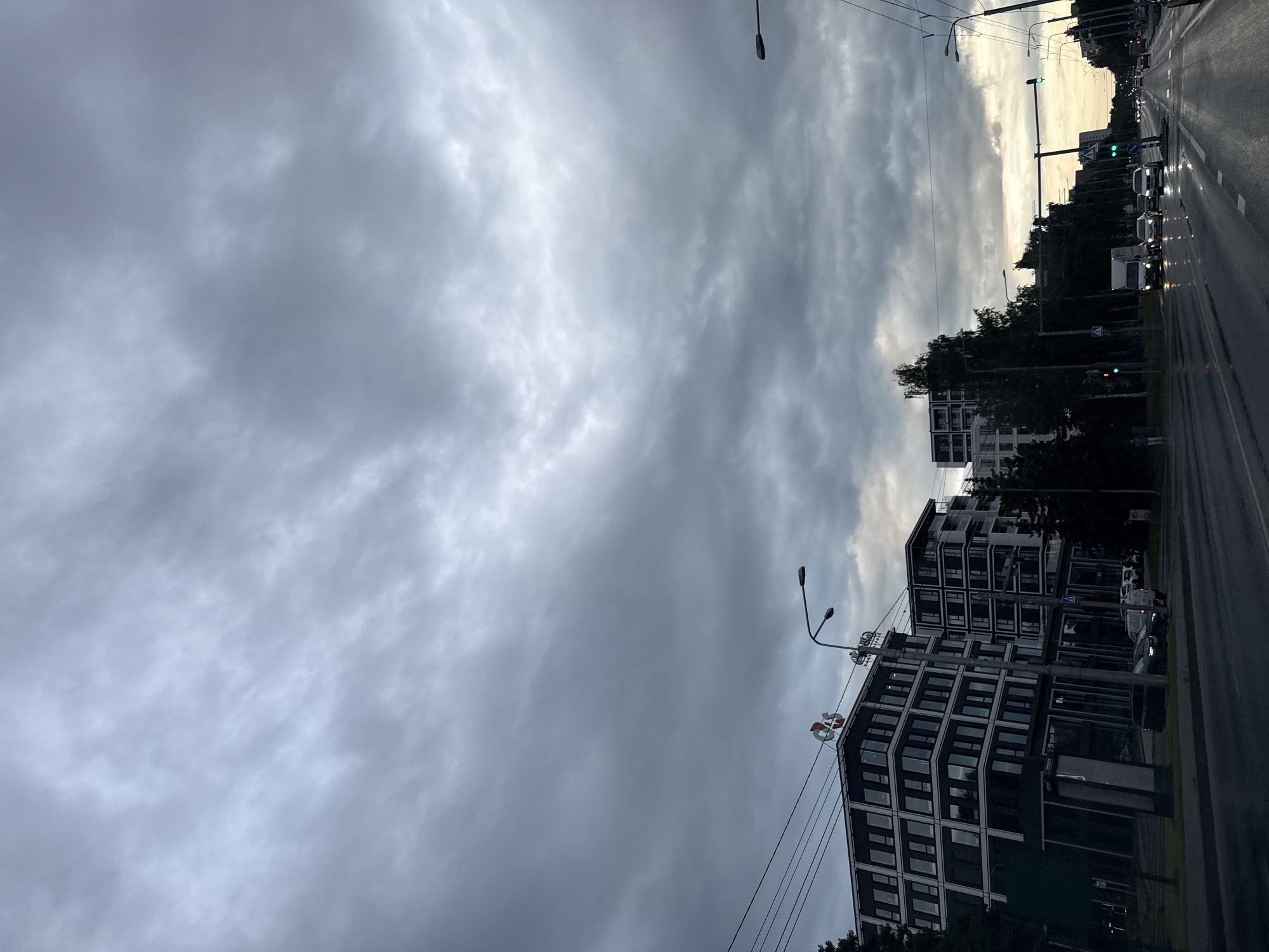 The image depicts a street scene in Tallinn, Estonia, under a dramatic, overcast sky. The perspective is from a vehicle on a wet asphalt road, suggesting recent rain or current drizzle. On the left, modern apartment or office buildings with dark facades and numerous windows are visible. One building has a red and white logo at the top. On the right side of the road, trees line the street, interspersed with more modern buildings in the distance. Traffic lights are visible, with some showing green. Several vehicles are on the road, including cars, vans, and a truck, some with headlights on. Overhead power lines and streetlights are prominent features. The overall atmosphere is somber and slightly moody due to the dark clouds and muted light.