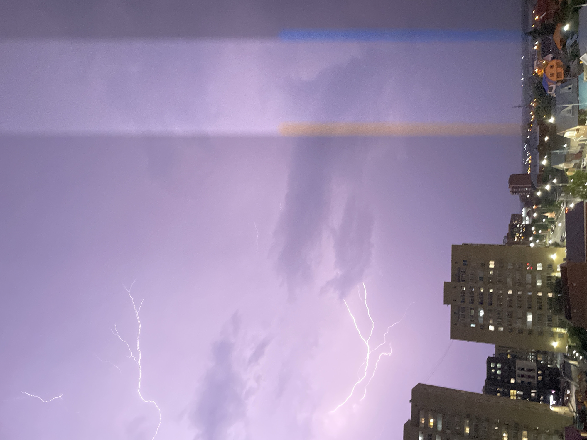 A lightning strike illuminates the purple-hued night sky over Petropavl, Kazakhstan. The image captures a cityscape dominated by apartment buildings, their windows glowing with artificial light. The foreground is occupied by the upper portions of two tall apartment complexes, casting shadows over a street lined with houses and streetlights. In the distance, the city stretches out with more buildings and lights, suggesting a suburban or semi-urban environment. Two distinct vertical bands of color, one orange and one blue, appear in the sky, possibly a reflection or an artifact of the camera.