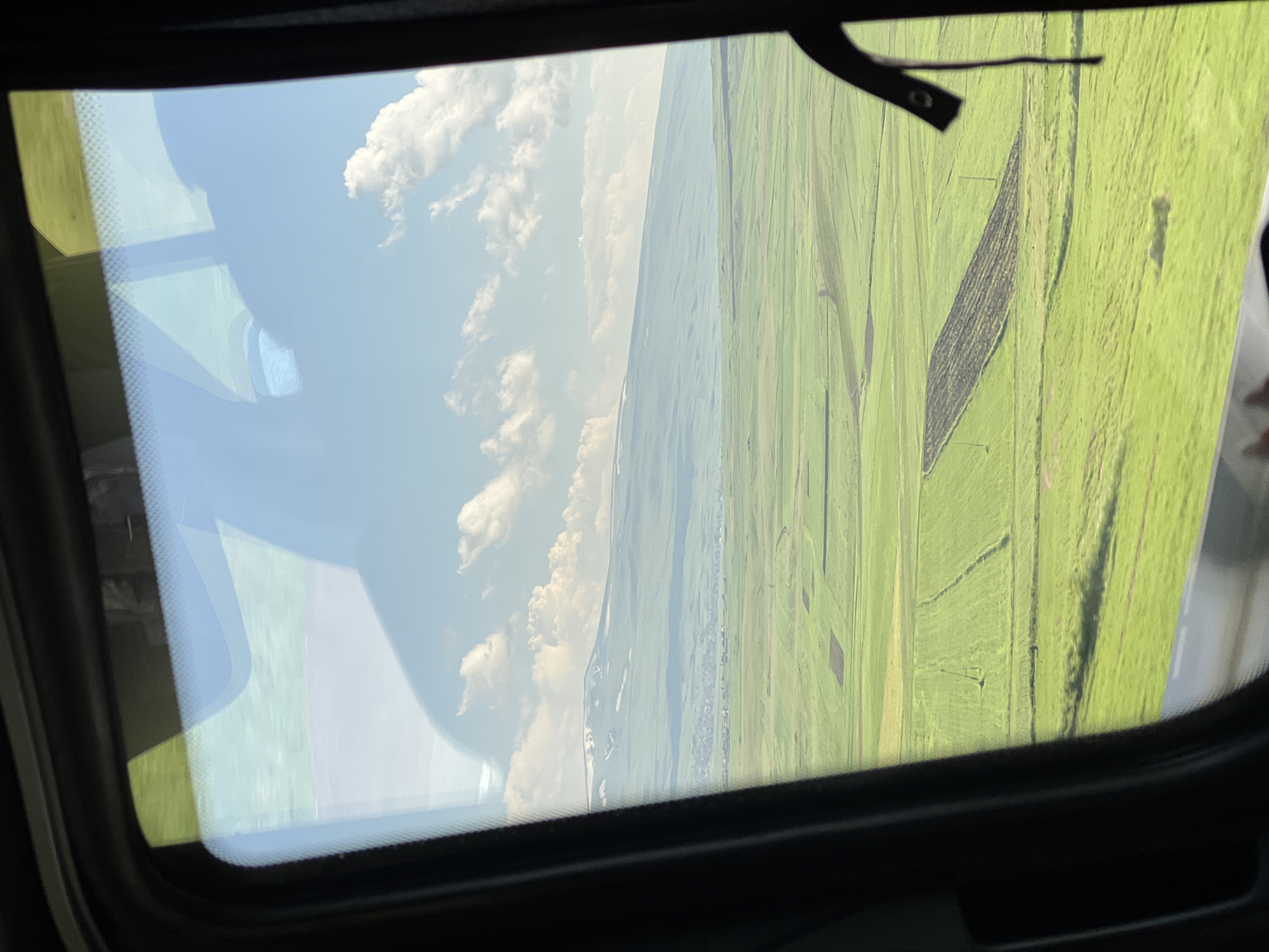 This media file is a photograph taken from inside a vehicle, likely a car or van, looking out through the windshield. The primary subject is a vast, green landscape divided into rectangular fields, suggesting agricultural land. The fields are predominantly a vibrant green, with some areas appearing darker, possibly indicating different crops or soil conditions. One large, dark rectangular patch in the middle of the green fields stands out.
The sky above is blue with scattered white, fluffy clouds. The perspective indicates that the photograph was taken from a moving vehicle, as suggested by the reflections on the windshield, which show parts of the vehicle's interior, including a dark frame around the windshield and a black strap or lever. These reflections create a layered effect, obscuring some of the view of the landscape.
The scene appears to be set during the daytime, with bright sunlight illuminating the landscape. The time of day seems to be late morning or early afternoon, given the brightness and the angle of the sun. The weather appears clear and sunny, with fair-weather clouds. The landscape suggests a rural or semi-rural location, characterized by open fields and a distant horizon.
There are no people or explicit events depicted. The overall impression is one of a scenic drive through the countryside. The image is oriented vertically, suggesting it might have been taken while tilting the camera or during a turn. There is no visible text within the image. The location context provided is Tsilkar, Armenia, which aligns with the appearance of the agricultural landscape.