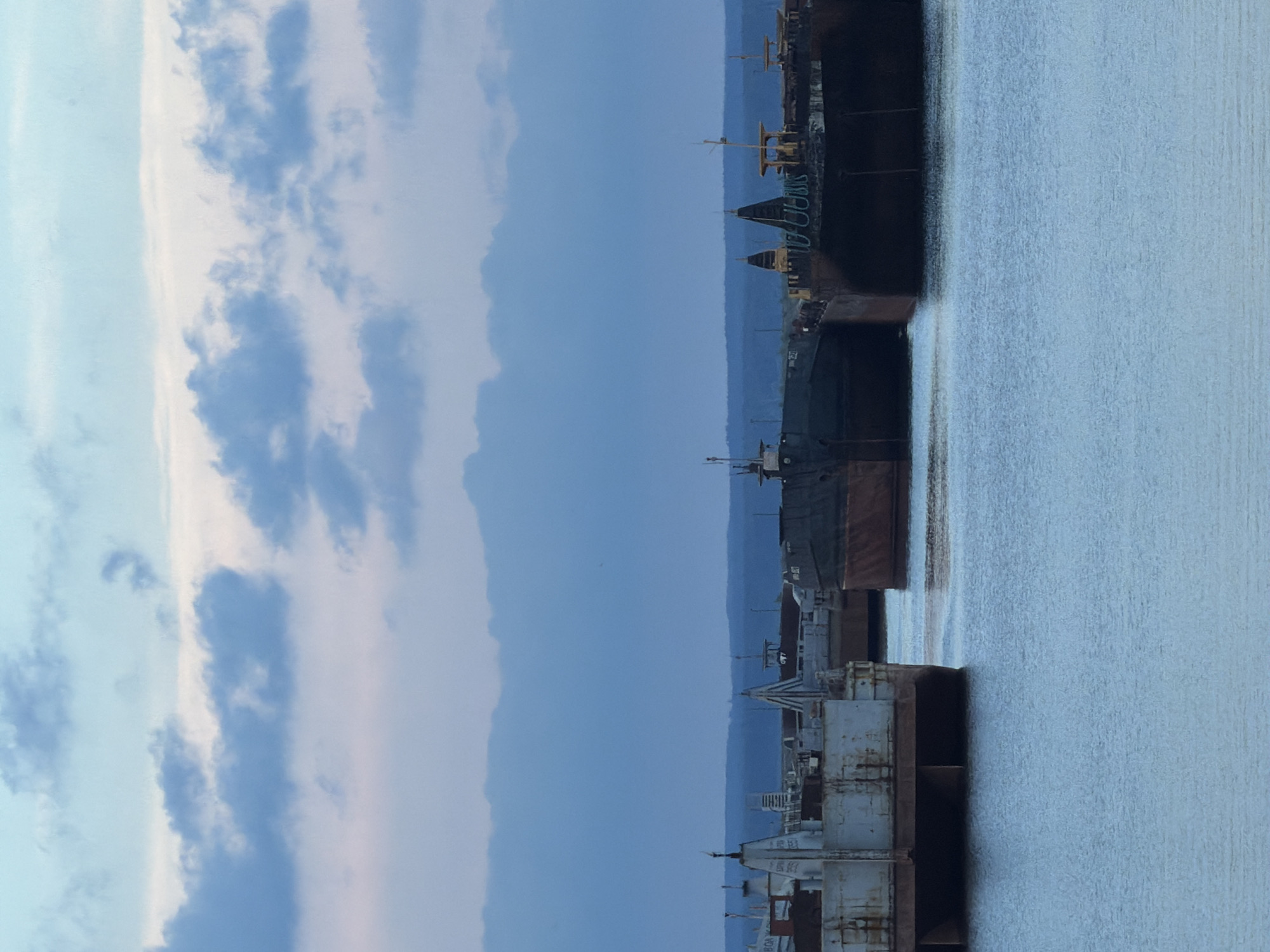 The image shows a body of water with several large, industrial barges moored. The sky above is a pale blue with scattered clouds. The barges appear to be docked at a port or industrial area. The scene is calm and still, with no people visible. The overall impression is one of industrial activity or storage.