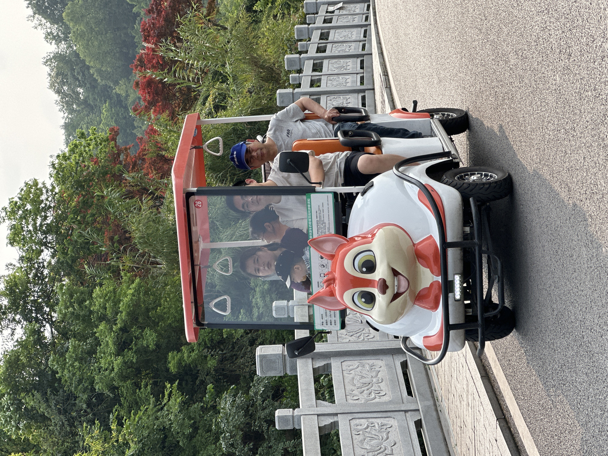 Two adult males are seated in a white and orange golf cart with a cartoon squirrel face attached to the front. The man in the front seat is wearing a blue baseball cap. The golf cart is on a paved surface, and a stone railing is adjacent to it. Lush green trees and vegetation are visible in the background. The location is Liangjiang, China.