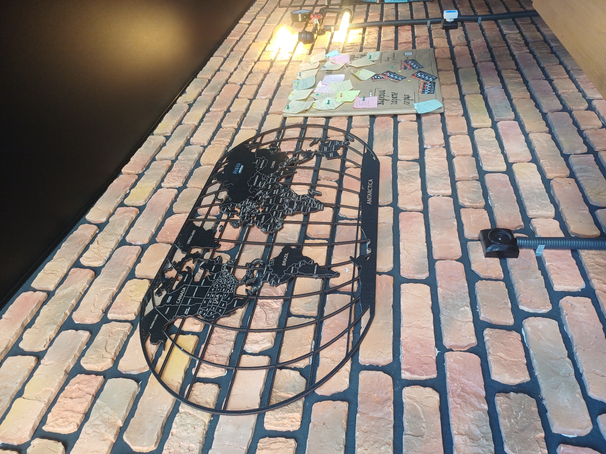 The image depicts a brick wall in what appears to be an indoor establishment. A large, black, metal cutout of a world map is mounted on the wall. To the right of the map, a corkboard is covered with numerous sticky notes and papers, attached with pins. Some of these notes have handwritten text. Industrial-style lighting fixtures are visible on the right side of the image, one of which is a bare bulb emitting light. A black security camera is mounted on a flexible conduit below the world map. The brickwork is varied in color, with shades of orange, yellow, and pink, and the mortar is dark gray. The ceiling above the wall is black. The scene is static and does not contain any people or visible actions. The location is identified as Baryshivka, Ukraine.