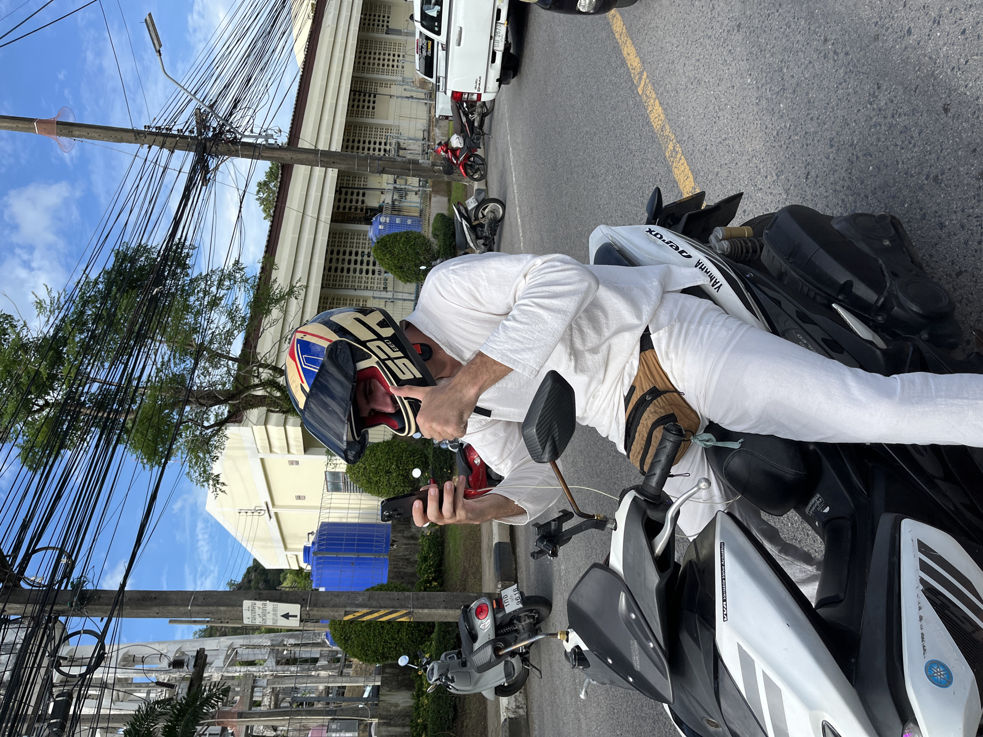 A man wearing a white tunic, white pants, and a motorcycle helmet is seated on a scooter in an outdoor urban environment. He is holding a black smartphone in his left hand, pointing it forward, and giving a thumbs-up with his right hand. The scooter has a black and white body, with a fanny pack around the man's waist. In the background, there are other scooters, a white pickup truck, buildings, trees, utility poles with numerous wires, and a blue water tank. A street sign with Thai script and an arrow pointing up is visible on a pole. The scene appears to be on a street in Phuket City Municipality, Thailand, given the presence of Thai script and the overall tropical urban setting.