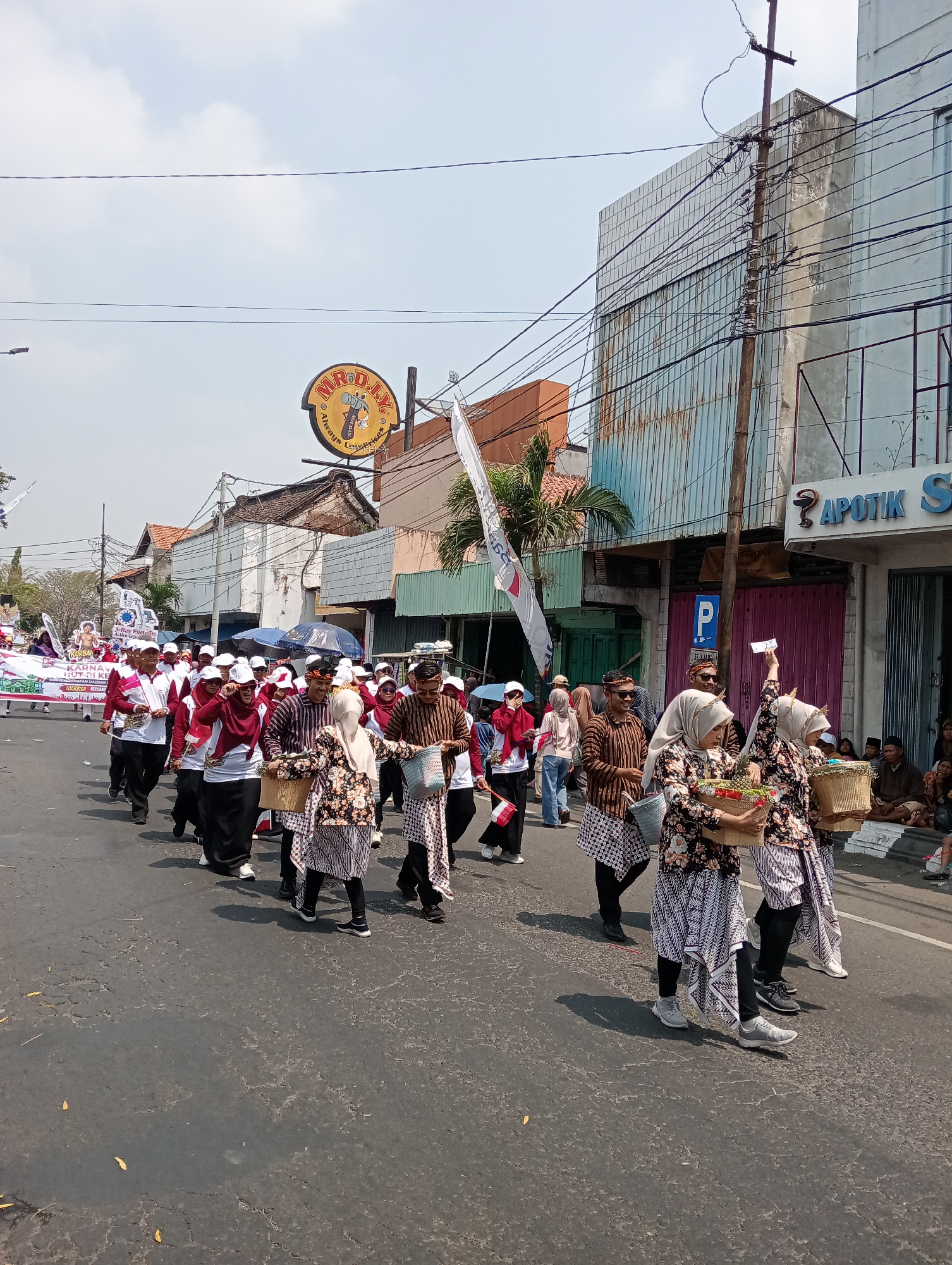 In a procession on a street in Demak, Indonesia, a group of people are walking. The participants are wearing traditional Indonesian clothing, including batik sarongs and patterned tops. Some individuals are carrying baskets filled with produce, such as fruits and vegetables. Others are holding flags or banners. The street is lined with buildings, some of which have a faded, aged appearance, suggesting the presence of older architecture in the city. A sign for "MR. D.I.Y." is visible, indicating a commercial establishment nearby. The weather appears to be sunny with a clear sky. The overall scene depicts a cultural event or parade taking place in a bustling urban environment.