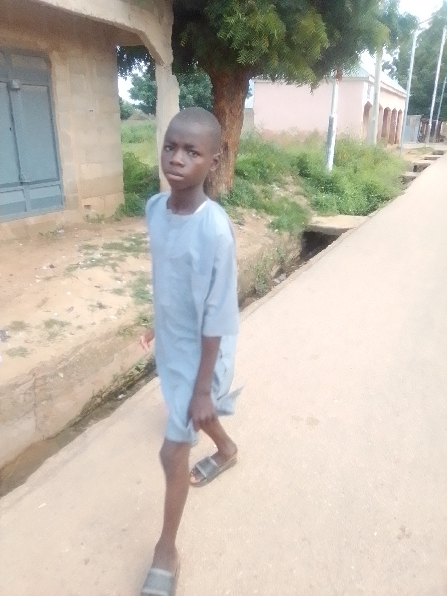 A young boy, appearing to be of African descent and approximately 8-10 years old, walks on a paved pathway. He is wearing a light blue, loose-fitting garment and dark-colored sandals. The boy's gaze is directed forward, slightly to his left. The environment appears to be an outdoor setting in a village or rural area, possibly in Liji Hill, Nigeria, with a dirt path or drainage area to his left and a paved road to his right. In the background, there is a building constructed with concrete blocks and a light pink building. Vegetation, including trees and grass, is visible throughout the scene. The lighting suggests it is daytime.