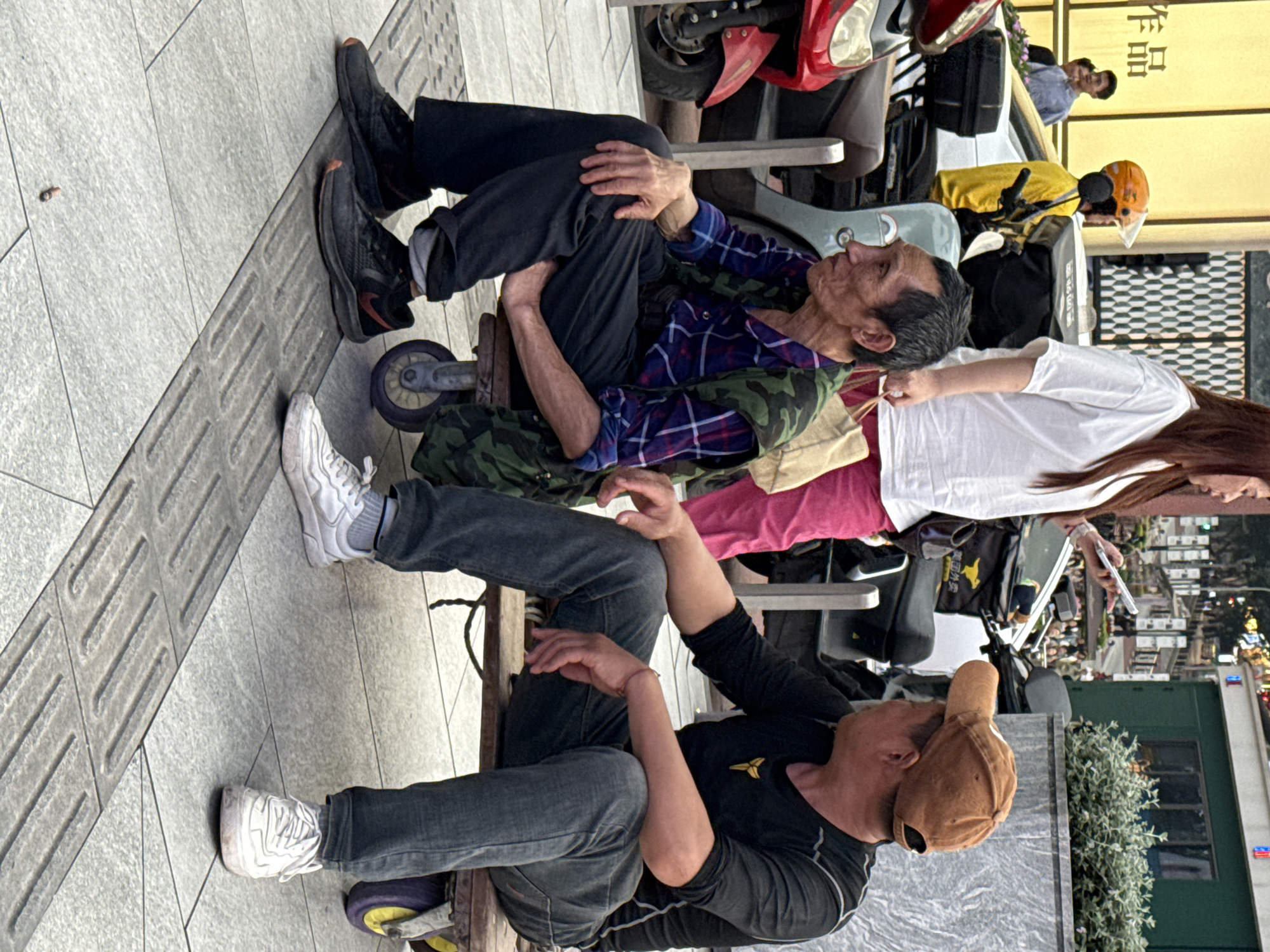 Two men, one older and one younger, are seated in makeshift wheelchairs on a city sidewalk. The older man wears a plaid shirt, camouflage vest, and dark pants with black shoes. The younger man is dressed in a black shirt, jeans, a brown cap, and white sneakers. They appear to be looking in different directions, with the younger man seemingly raising his hand. A woman in a white shirt and pink pants stands nearby, possibly interacting with them.
The sidewalk is tiled and textured. Behind them, there is a variety of objects, including a scooter, a food delivery vehicle with a yellow helmet, luggage, and other parked scooters. In the background, there are more people, buildings, a bush, and a street with lights, suggesting an urban environment.
The photo was taken in the city of Liangjiang, China.