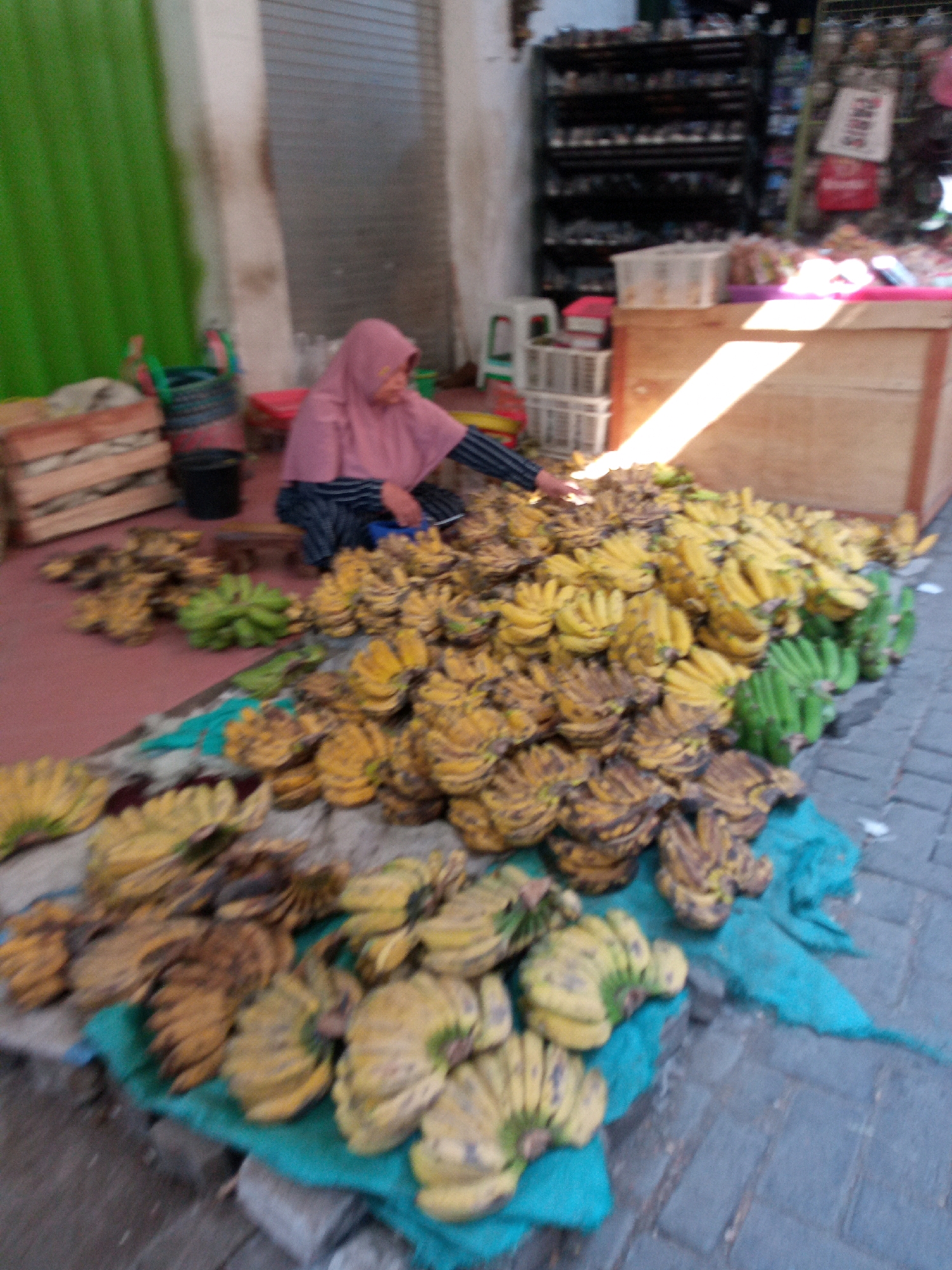 A vendor in Balun, Indonesia, sits behind a large display of bananas. The bananas are in various stages of ripeness, with some green and some yellow with brown spots. The vendor, wearing a pink hijab and a striped shirt, is seated on the ground, her hands resting on the fruit. Behind her, there are wooden crates, plastic baskets, and a shelf stocked with goods. The scene appears to be an outdoor market stall, with cobblestone paving visible in the foreground.