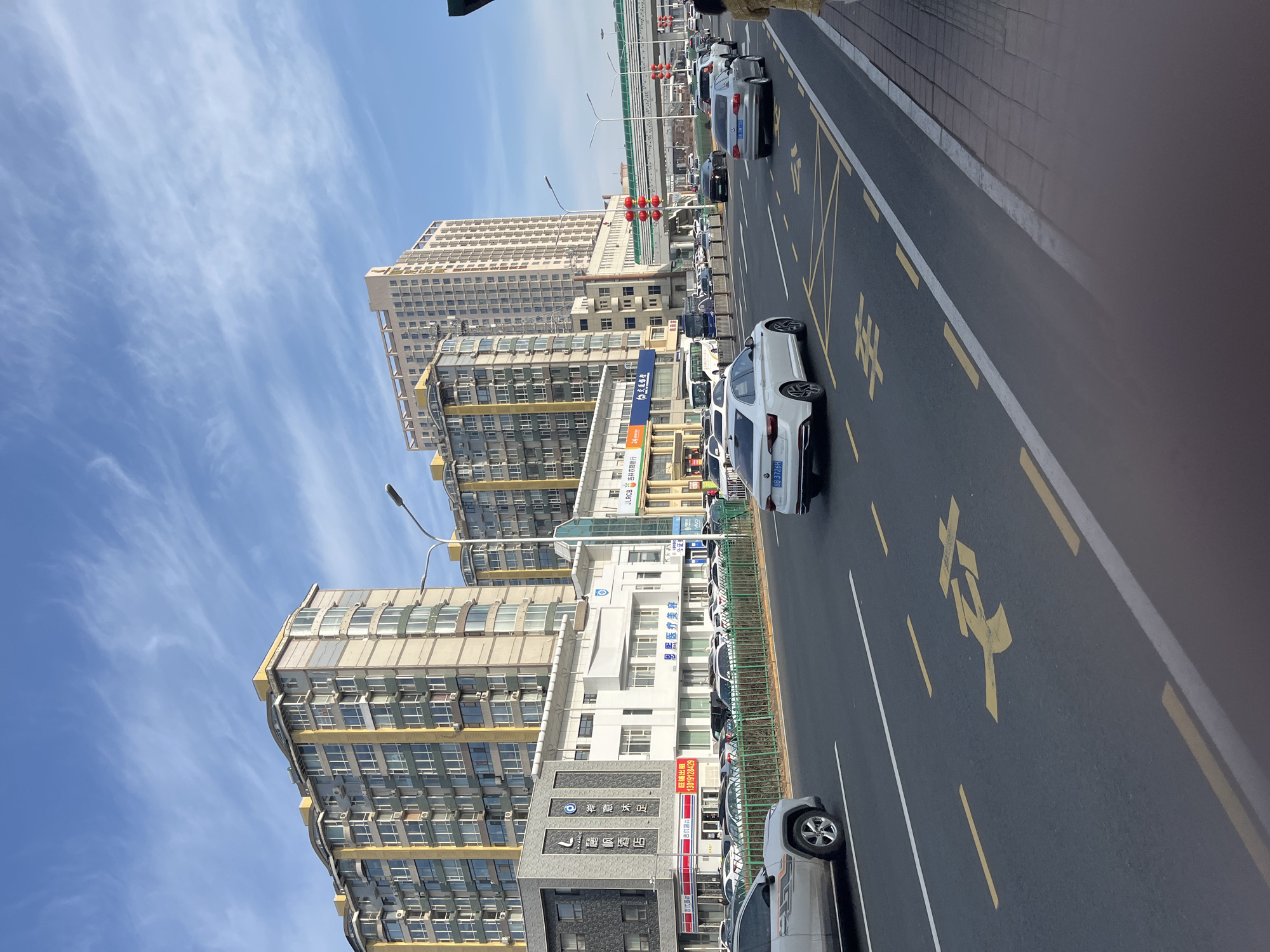 The image depicts a street scene in Changchun City, China, under a blue sky with scattered clouds. Tall, multi-story buildings line the left side of the street, showing residential and commercial establishments. There are several businesses visible, including "Lavande Hotel", a medical aesthetics clinic, and a branch of the Jilin Rural Commercial Bank. A street-level view reveals a line of cars parked along the curb, with several other vehicles moving along the road. The road markings are in yellow, with characters painted on the pavement. A person is partially visible at the very top right corner of the frame. A bridge or overpass is visible further up the road. Traffic lights are visible in the middle ground.