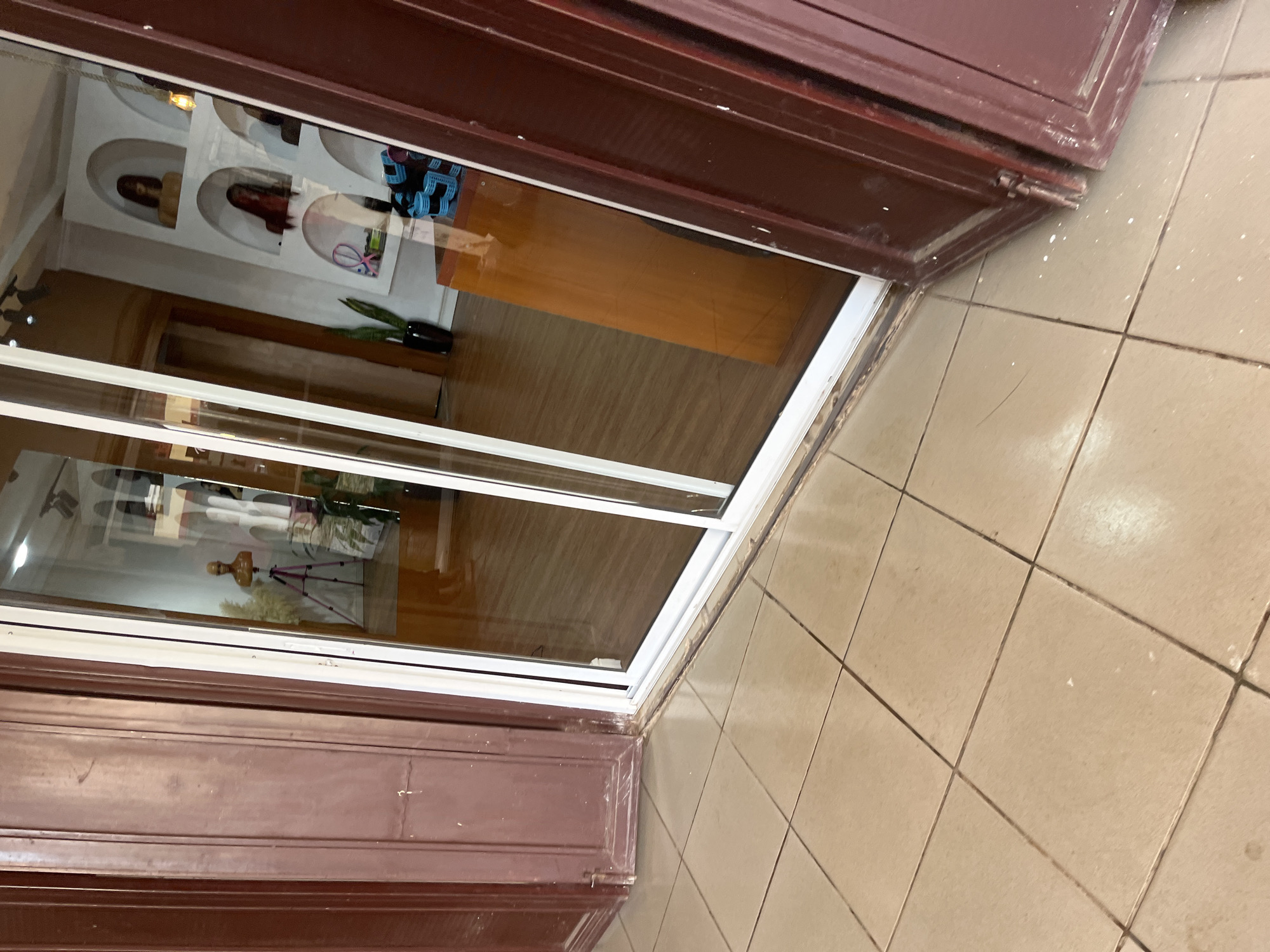 The image depicts the exterior of a shop with a glass sliding door. The foreground shows a tiled floor that appears worn and possibly dirty in places. To the left, a dark brown wooden door with vertical panels is visible, showing signs of age and wear. To the right and through the glass door, the interior of the shop is visible.
Inside the shop, the floor is wooden. There are shelves on the wall displaying mannequins with wigs. One mannequin on a tripod is visible to the left of the display shelves. To the right, a brown wooden counter or display unit is present. Blue hair curlers are also visible on a shelf. A black vase with a plant is to the left of the shelves. The lighting inside the shop suggests it is daytime. The overall impression is of a hair salon or wig shop.
While the image shows the exterior of a shop, it does not provide any specific visual cues to pinpoint its exact location within the city of Gura Top, Nigeria.