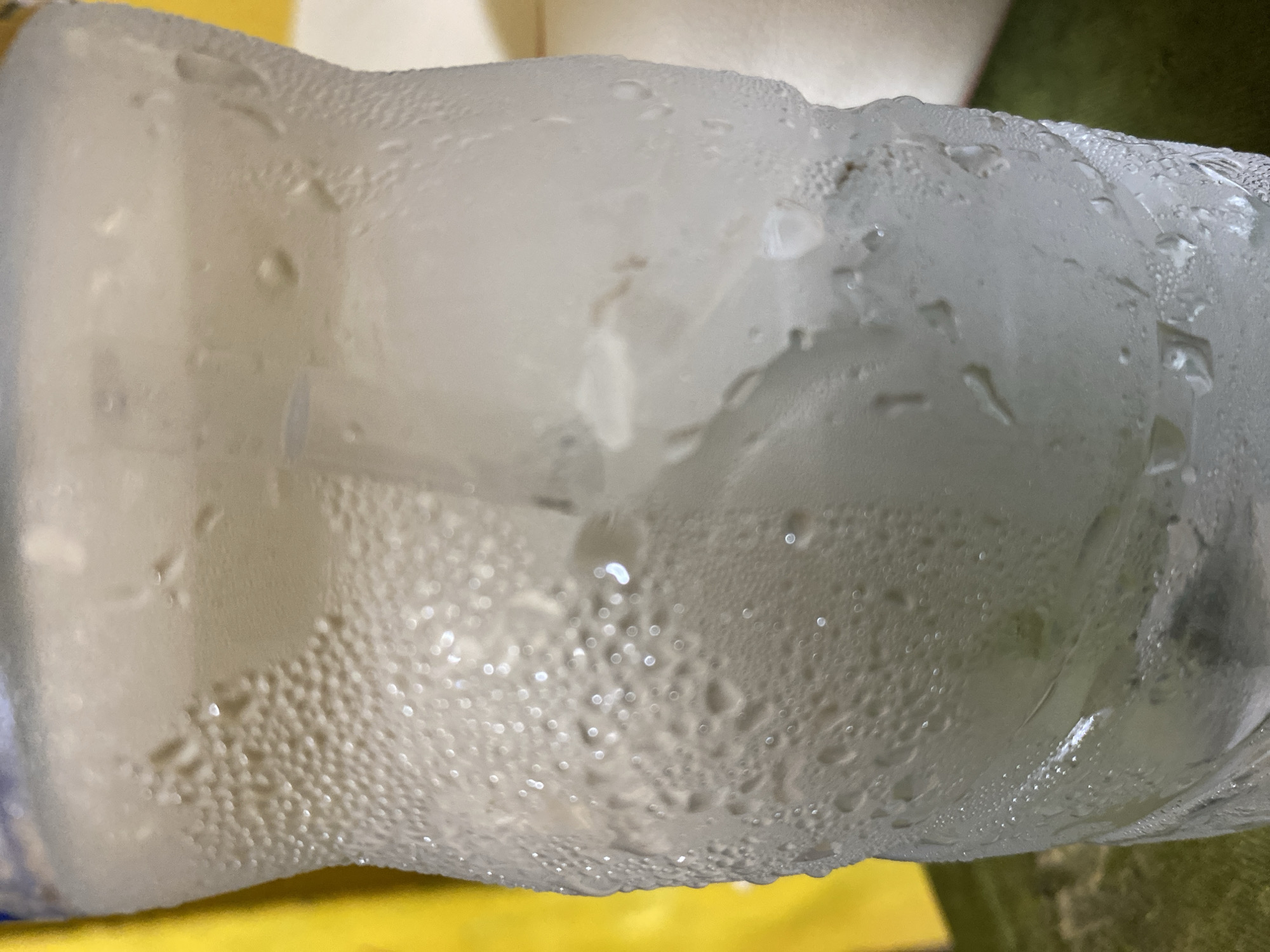 The image shows a plastic bottle covered in condensation droplets, likely from a cold beverage. The bottle appears to be slightly deformed and contains a liquid inside. The background is blurred and includes yellow and green elements. The location is unknown, with no information in the image to indicate it is in Tanjongkapai, Indonesia. There are no people visible in the image.