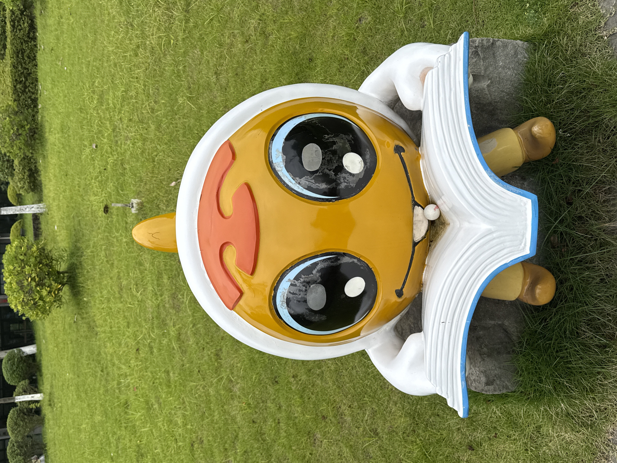 A statue of a yellow, cartoonish character wearing a white and orange hood sits on a grey rock in a grassy area. The character has large blue and black eyes with white pupils and a small white ball in its mouth. It is holding a white, open book with blue trim. The background consists of green grass, some bushes, and white posts. The scene is outdoors, possibly in a park or public space in Jiangjin, China.