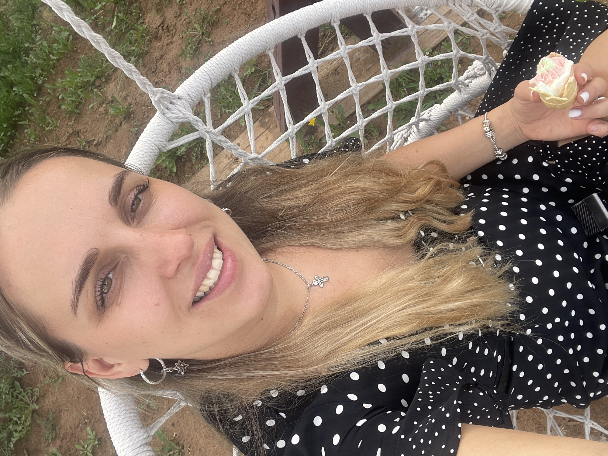 A woman with blonde hair is reclining in a white rope hammock, holding an ice cream cone in her right hand. She is smiling at the camera. She is wearing a black and white polka dot dress. She has silver hoop earrings with a floral design, a silver cross necklace, and a silver bracelet. The ground is visible through the hammock's netting. The location is implied to be in the city of Chaikovsky, Russia.