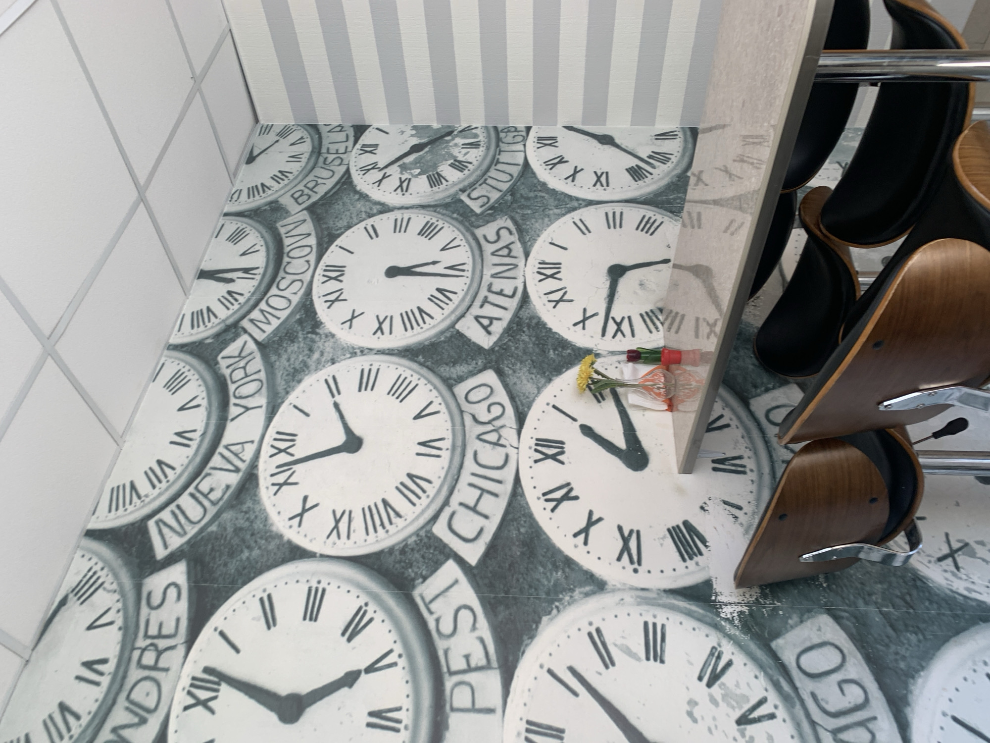 The image shows a close-up, floor-level view of a decorative wallpaper featuring numerous clock faces with Roman numerals, each labeled with a city name. The cities visible include BRUSSELS, MOSCOW, NEW YORK, STUTTGART, ATHENS, CHICAGO, and PEST. The clocks have varying times displayed. The wallpaper is on the floor, and the perspective is unusual, making the clocks appear as if they are on a wall or a flat surface that is tilted. Above the wallpaper, a wall with light gray and white vertical stripes is visible, along with a white tiled wall on the left. A metal rack holding several black and wooden chairs is partially visible on the right side of the image. The chairs are stacked and positioned in a way that suggests they are for seating in a waiting area or a salon. A small bouquet of flowers and some colorful items, possibly decorations or toiletries, are placed on top of the wallpaper near some of the clocks. The lighting appears to be artificial, creating a slightly reflective surface on the wallpaper. The overall impression is of a stylish and eclectic interior design, possibly a waiting room or a themed establishment. There are no people visible in the image.