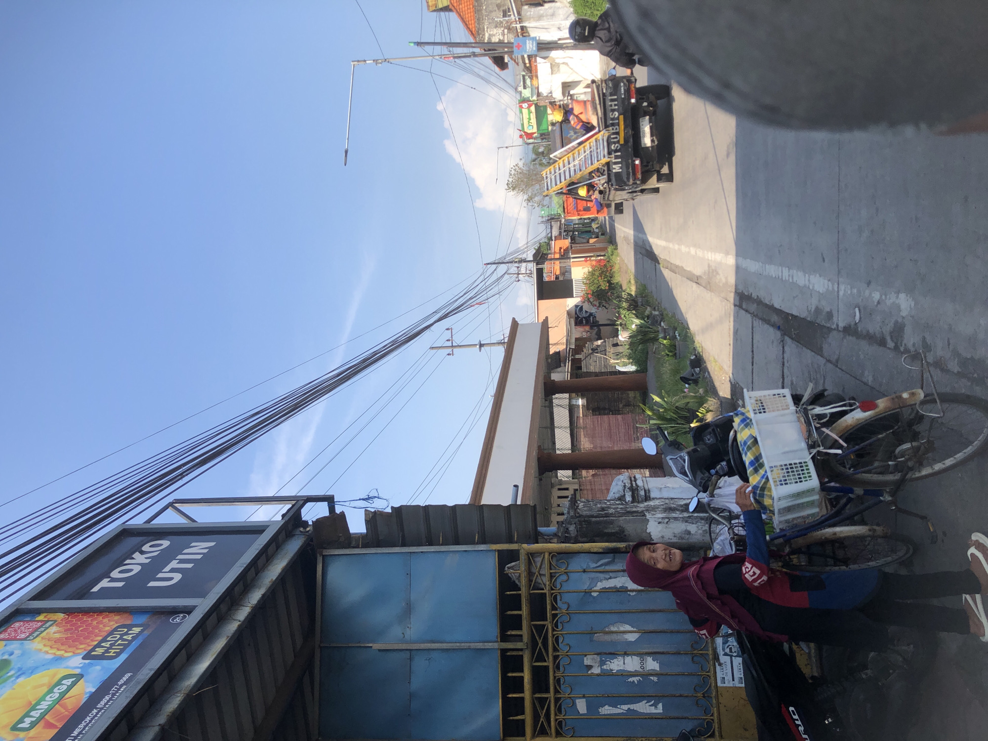 In Modong, Indonesia, a street scene unfolds under a clear blue sky. A woman, wearing a maroon headscarf and a dark-colored shirt with red accents over black pants, stands beside a bicycle. The bicycle has a white basket on the front, filled with yellow and checkered fabric. A black motorcycle is parked next to the woman and the bicycle.
Behind them, a building with a blue metal gate and a sign reading "TOKO UTIN" is visible. The gate has some peeling stickers. A yellow metal fence borders the gate area. Further down the street, a white building with pillars stands, and beyond that, a row of buildings line the road.
A dark-colored Mitsubishi pickup truck, carrying a ladder and at least two individuals wearing orange vests and hard hats, drives down the street. Another motorcycle, with a rider wearing a black helmet, is in front of the truck. The road is paved with white lines marking the lanes. The environment appears to be a typical urban or suburban street in Indonesia.