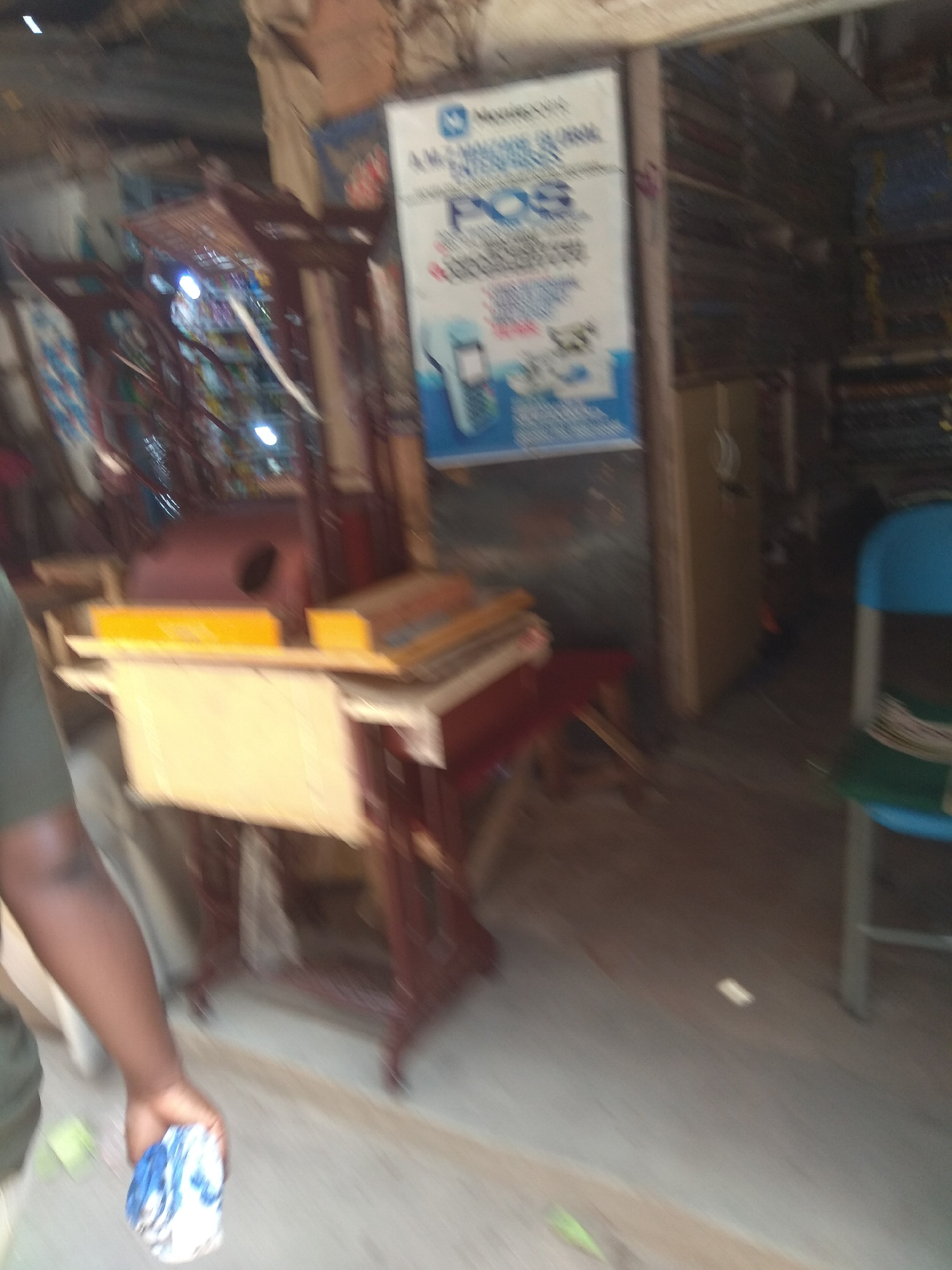 In a market stall in Jalingo, Nigeria, a person's arm and hand are visible, holding a blue and white patterned cloth. The stall appears to be selling various goods, including what looks like fabric rolls and a POS machine advertisement. A blue chair is also visible to the right of the image. The scene suggests a commercial environment.