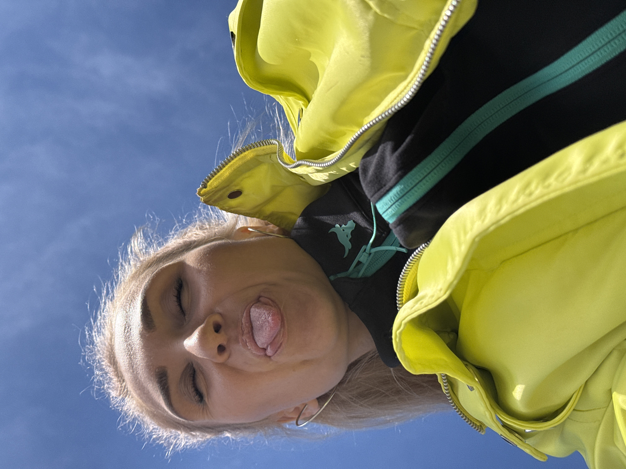 The image shows a young woman captured from a low-angle perspective, looking down towards the camera against a clear, bright blue sky. She has blonde hair and fair skin. She is wearing a vibrant yellow or neon-green puffer jacket with a visible silver zipper. Beneath the jacket, she wears a black garment, possibly a hoodie, which features a prominent teal zipper and a light blue or teal graphic symbol on the chest area. Small hoop earrings are visible on her ears.
The woman is making a playful or silly face, with her eyes closed, cheeks pushed upwards, and her tongue sticking out. This conveys a humorous and lighthearted emotion. The background is a vast, cloudless blue sky, indicating a sunny outdoor setting during daylight hours. The strong sunlight illuminates her features and clothing. The image was taken in Chaikovsky, Russia. There is no visible text.