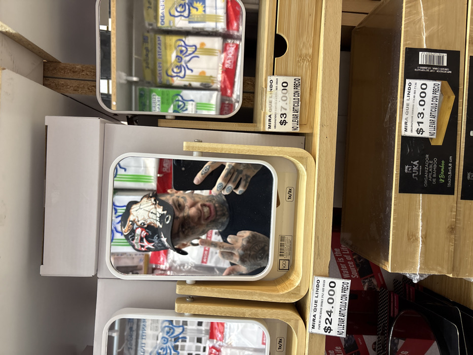 The image displays a mirror selfie taken by a male individual within a "Mira Que Lindo" retail store in Buenos Aires, Argentina. The subject is reflected in a rectangular vanity mirror with a light wooden frame and stand, prominently featuring extensive black tattoos on his face, neck, and hands, along with large earlobe gauges. He wears a black baseball cap with a multi-colored design and a black t-shirt. The individual holds a black smartphone in his left hand, positioned to capture the reflection. His right hand is raised with the middle finger extended, and his facial expression is a grimace with an open mouth, exposing his teeth.
The mirror is part of a retail display on a white shelf, bearing a price tag of "$24.000" (Argentine Pesos) for an "ESPEJO TOCADOR DOBLE." Adjacent to it on the right is another display mirror with a small drawer, priced at "$37.000." Both price tags include the phrase "NO LLEVAR ARTICULO CON PRECIO" (Do not take item with price). Reflections in the surrounding mirrors show packaged consumer goods, possibly toiletries, featuring a blue bow design. On the shelf directly below, stacked "JUKA HOME style" bamboo organizers are visible, one with a price tag of "$13.000." The environment is consistent with an indoor retail space, characterized by white shelving units and various consumer products.