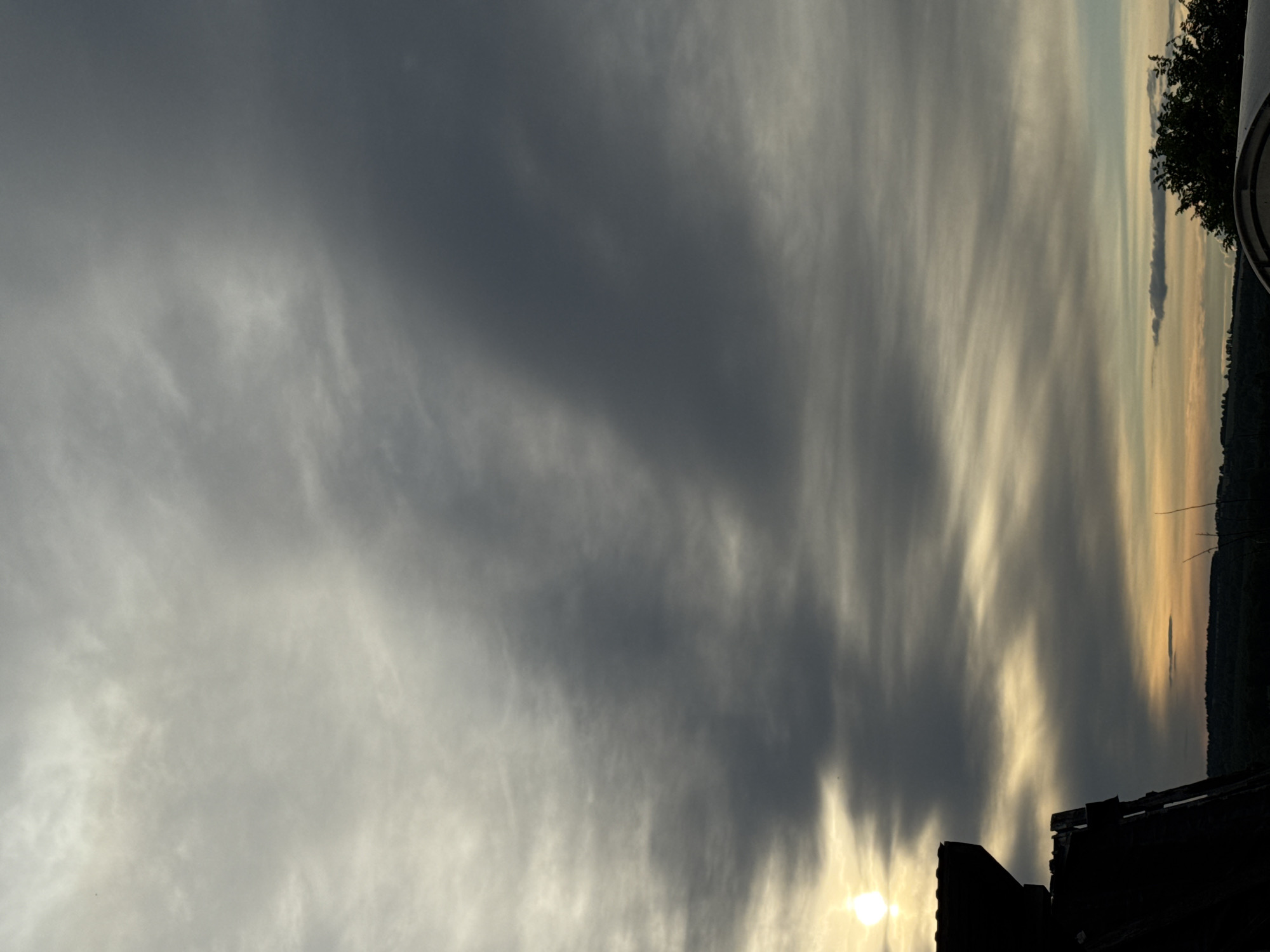 The image captures a dramatic sky with dark, heavy clouds dominating the view. Streaks of lighter, yellowish light break through the cloud cover, suggesting either sunrise or sunset. At the bottom of the frame, a dark, silhouetted rooftop is visible. On the right side, a strip of land with trees can be seen in the distance, potentially indicating a horizon line or a body of water. The information provided does not offer adequate details to discern the location in Nizhnyaya Gar', Russia.