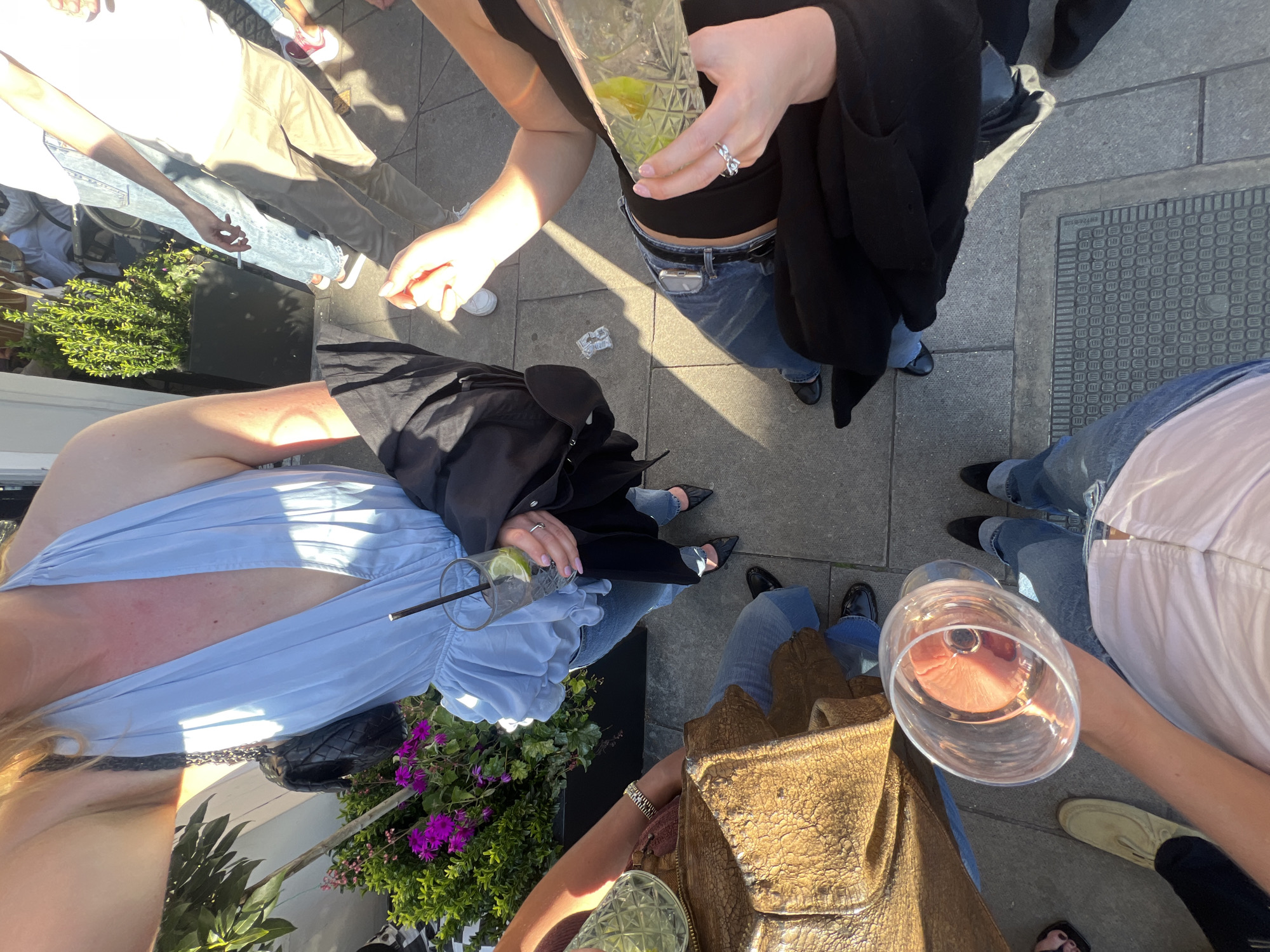 A top-down shot depicts a group of five individuals standing in a circle on a paved surface. Each person is holding a glass, suggesting a social gathering. The individuals are dressed in casual clothing including jeans, dresses, and blouses. Accessories such as rings, belts, and bags are visible. A planter with greenery and purple flowers is near one of the individuals. The setting appears to be outdoors, with shadows indicating daylight. The photograph lacks context for specifying its location.