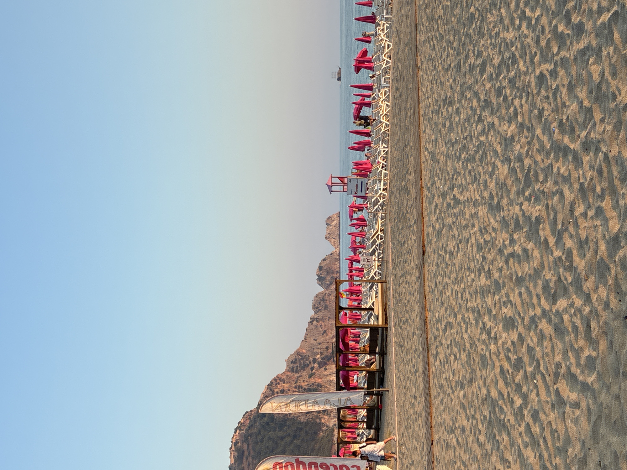 The image depicts a beach scene on a clear day in Alanya, Turkey, likely in the late afternoon, as evidenced by the warm lighting and long shadows. The foreground is dominated by textured sand. A wooden boardwalk runs horizontally across the mid-ground. Beyond the boardwalk, rows of white lounge chairs and bright pink umbrellas are neatly arranged, indicating a well-maintained beach resort or public beach area. Several wooden cabanas with thatched roofs provide shade.
In the background, the turquoise waters of the Mediterranean Sea stretch to the horizon, where a sailing ship is visible. The coastline is characterized by rugged, rocky mountains, a common topographical feature of Alanya. Two tall white flags with red lettering are planted near the left side of the image, partially obscuring the mountains behind them. A few people are visible on the beach, including two children walking near the flags and several individuals relaxing on lounge chairs or walking near the water. A lifeguard tower stands prominently in the middle ground, further suggesting a managed beach. The sky is a clear, pale blue, transitioning to a lighter hue near the horizon.