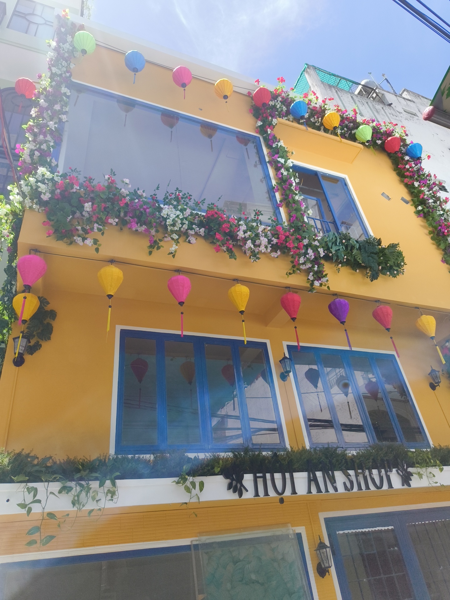 In Nha Trang, Vietnam, a vibrant yellow building is adorned with colorful floral arrangements and decorative lanterns. The facade features large blue-framed windows and balconies overflowing with blooming pink, white, and purple flowers, as well as lush green foliage. Strings of spherical lanterns in red, yellow, green, blue, and pink are suspended along the building's exterior, adding to its festive appearance. Below the upper windows, a white ledge showcases more greenery, and the text "HOI AN SHOP" is prominently displayed in black lettering. The building is illuminated by natural daylight under a clear blue sky, suggesting a bright and pleasant day.