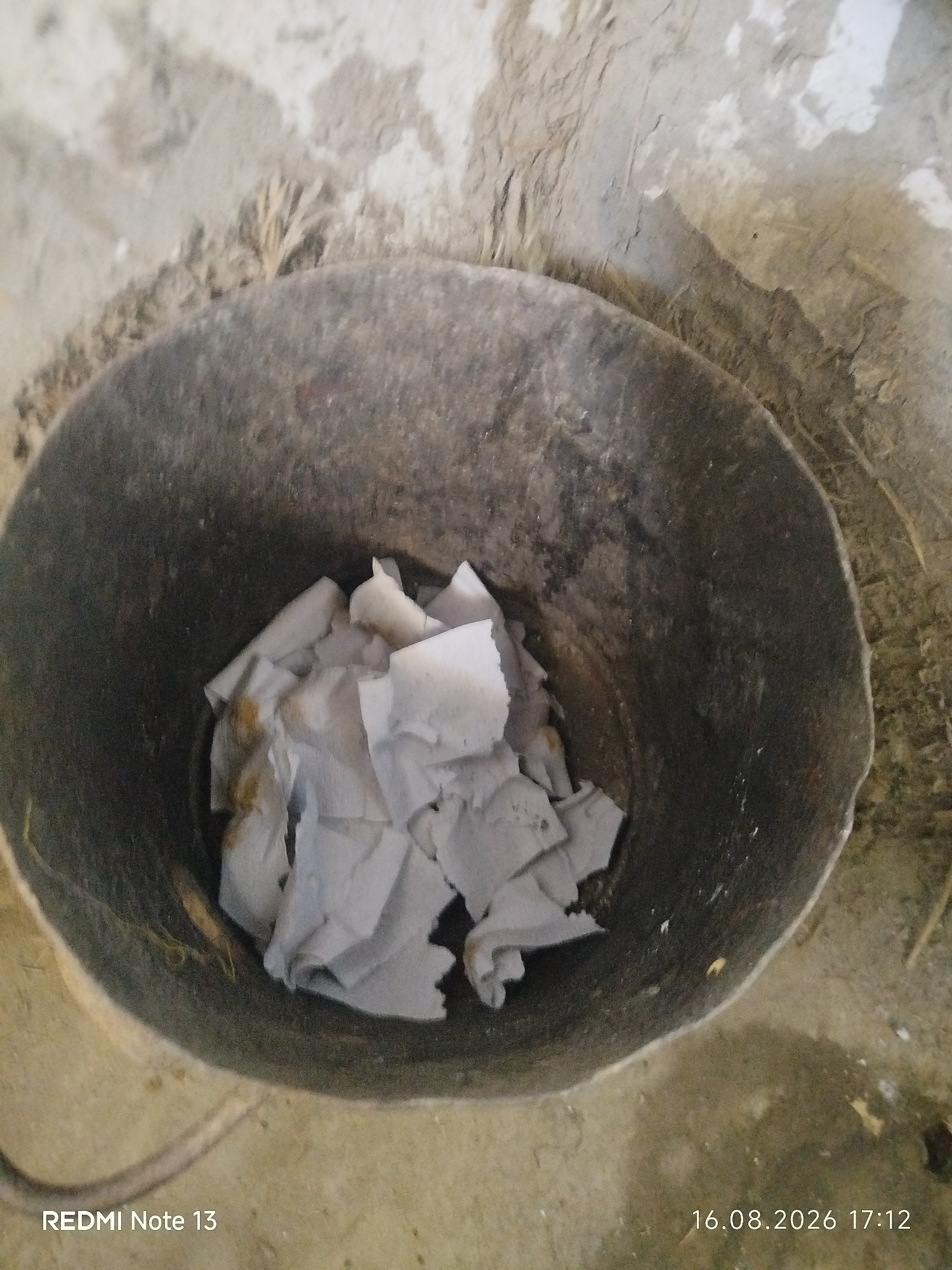 A close-up, overhead view captures a dark, weathered bucket filled with crumpled white paper. The bucket rests on a rough, sandy surface, with hints of dry vegetation and a light-colored, uneven ground surrounding it. The overall scene appears to be outdoors, with natural lighting. The timestamp "16.08.2026 17:12" and the text "REDMI Note 13" are visible in the lower right and left corners, respectively, indicating the image was taken with a Redmi Note 13 phone. The location is noted as Kungirat, Uzbekistan. There are no people or animals present in the image.