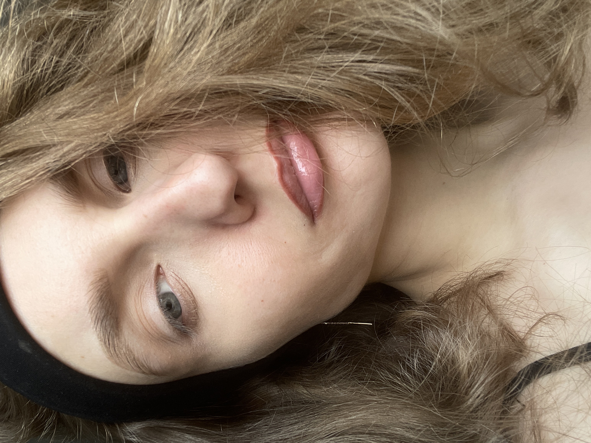 This close-up image, set in Podshivalovo, Russia, shows a person lying down, their head tilted or viewed from a unique angle, with significant portions of their face framed by their hair. The primary subject is a young individual with light brown, wavy hair that features blonde highlights, cascading around their head and partially obscuring their face.
Notable features include one visible eye with a light-colored iris (appears blue or grey) and natural-looking eyelashes. The eyebrow is well-defined, potentially shaped or microbladed. The lips are glossy and a pinkish-mauve color, slightly parted. The skin appears fair and smooth.
The most striking detail is a thin, metallic object, resembling an acupuncture needle or a very fine piece of jewelry, visibly inserted into the skin near the jawline/chin area, below the corner of the mouth. This suggests an activity or event related to a cosmetic or therapeutic procedure, such as acupuncture, dry needling, or a similar treatment. A black band, possibly a headband or part of clothing, is visible around the head on the left side of the frame.
The person's expression is neutral and calm, with no strong emotions discernible. The lighting suggests an indoor setting, and there is no visible text in the image.