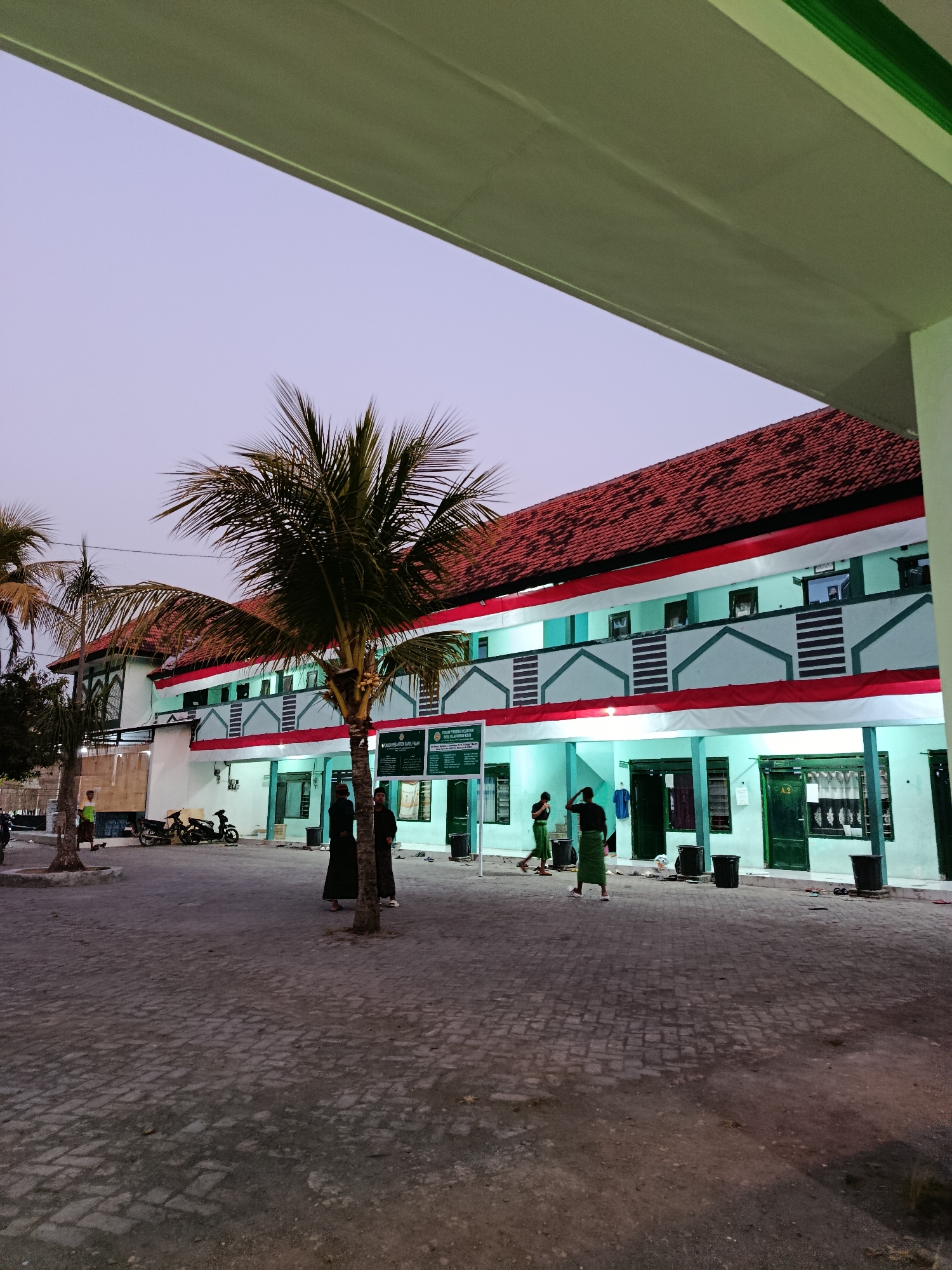 The image depicts the exterior of a two-story building in Ramban Wetan, Indonesia, possibly a school or dormitory, with a light blue facade, red tiled roof, and white and red decorative trim. Several palm trees are present, including one prominently in the foreground. The ground is paved with interlocking bricks. Two men in traditional Islamic attire are standing near the building, and two more are walking in the courtyard. A motorcycle is parked to the left. In the background, the sky is a pale purple, suggesting it is dusk or dawn. A green sign with Indonesian text is mounted on a pole near the entrance. The overall scene appears to be quiet and orderly.