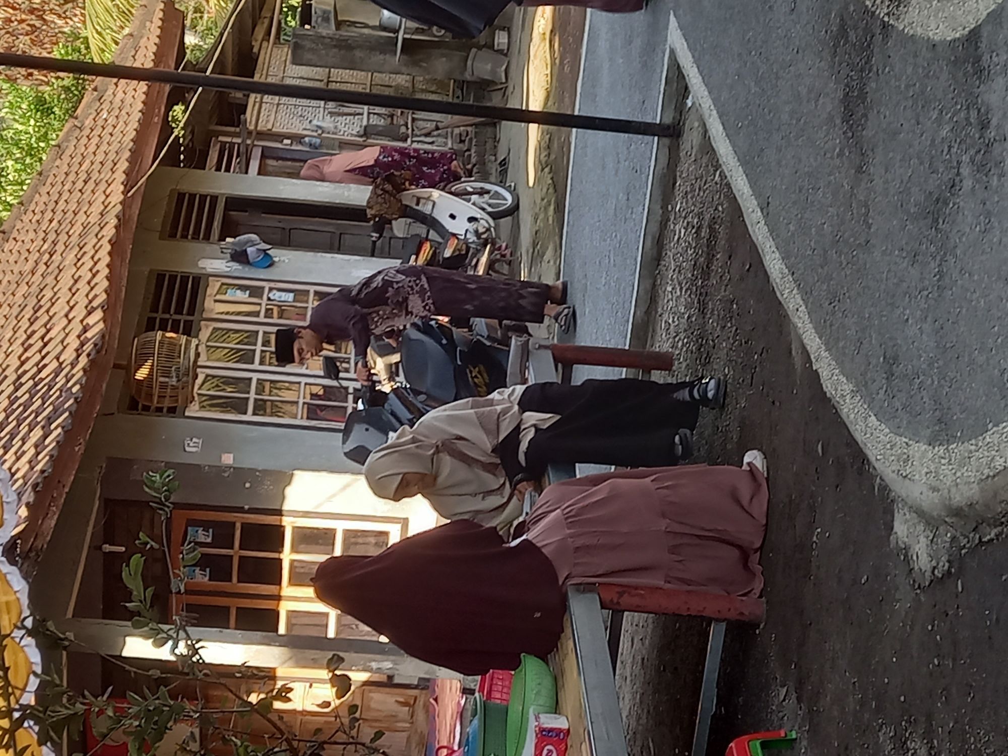 In the city of Tempos, Indonesia, two women are seated on a metal table next to a building with a tiled roof. One woman is wearing a dark maroon hijab and a light pink tiered skirt. The other woman wears a light beige hijab and a black skirt. Behind them, a man in a dark patterned shirt and pants is on a motorcycle, and another woman in a pink patterned dress stands near a doorway. The ground is a mix of dirt and asphalt.