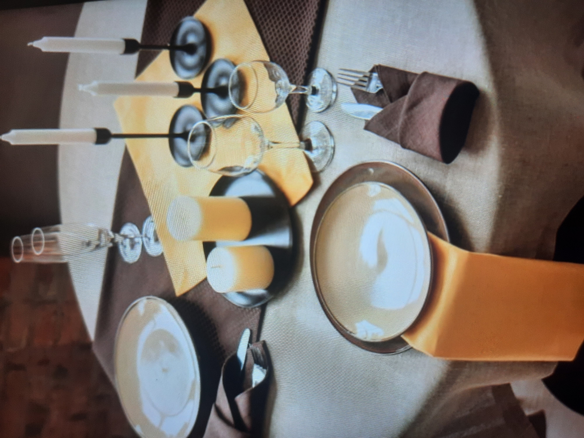 The image depicts a formal table setting for a meal. Two place settings are visible, each featuring a large white plate on a darker charger plate. One place setting includes a folded brown napkin with a fork and knife tucked inside. Various glassware, including champagne flutes and wine glasses, are also present. Three white taper candles in black candleholders are arranged on a golden placemat. The table is covered with a light-colored tablecloth, and the background shows a textured brick wall and a darker textured placemat. The scene appears to be indoors, possibly in a restaurant or dining room. The image does not provide any information about the city of Rosenheim, Germany, or any specific people.