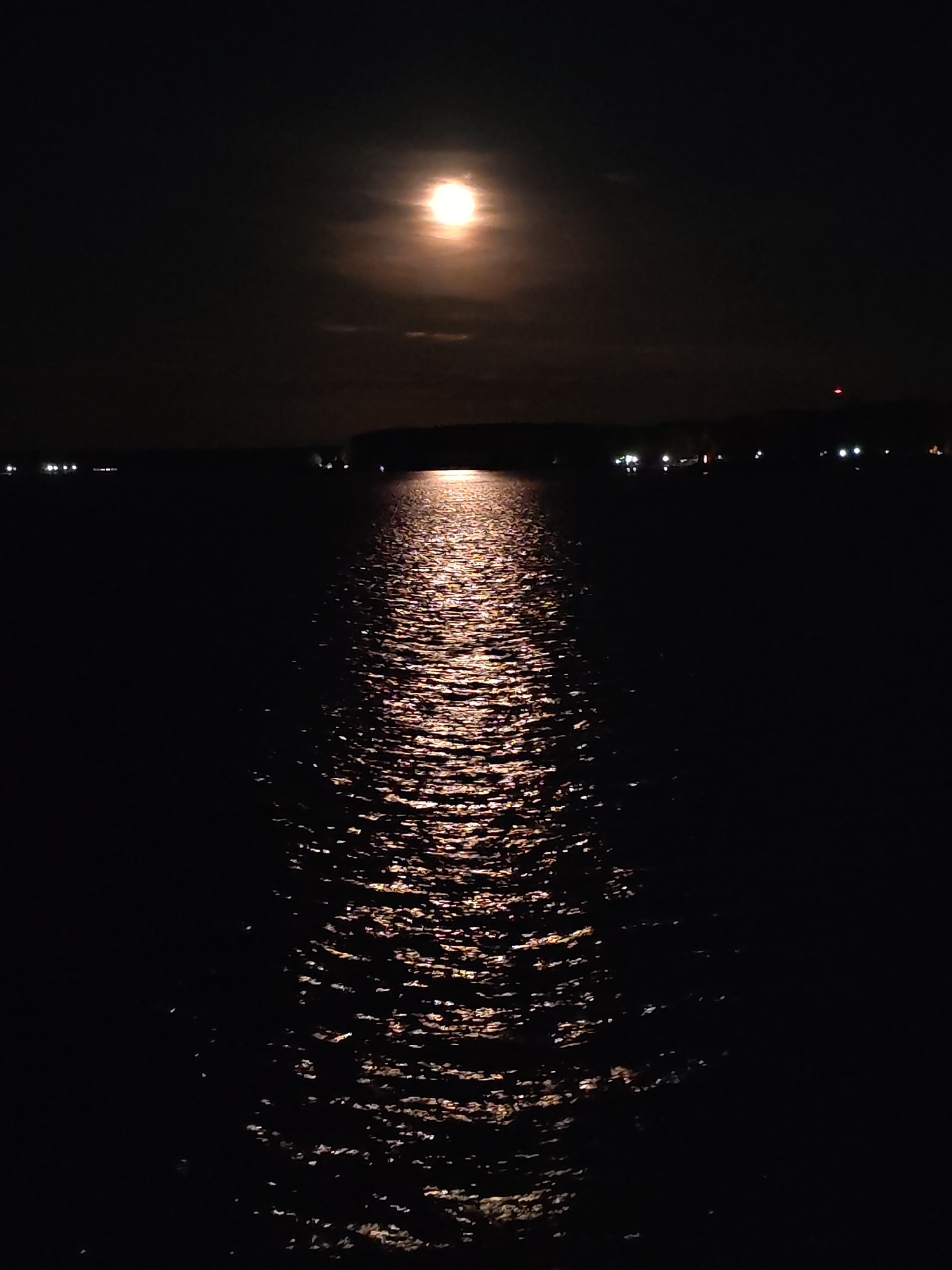 The image depicts a nighttime scene with the moon reflecting on a body of water, likely a lake or sea. The moon is visible at the top center of the frame, radiating light that creates a shimmering path across the water's surface. The surrounding environment is dark, with indistinct landmasses or city lights visible in the distance. The absence of specific landmarks or signage makes it impossible to pinpoint the location within the city of Unknown, Unknown.