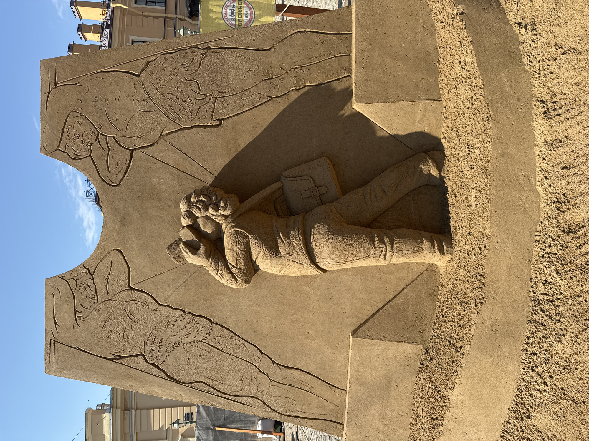 The image shows a large sand sculpture in Saint Petersburg, Russia. The sculpture features a central figure of a man lying down and reading a book, with a bag slung over his shoulder. The man is sculpted in three dimensions, while two other figures are depicted in relief on the background. These figures are also lying down, seemingly asleep or at rest. The sculpture is positioned outdoors, with buildings visible in the background. The sky is clear and blue. The overall environment suggests an urban setting.