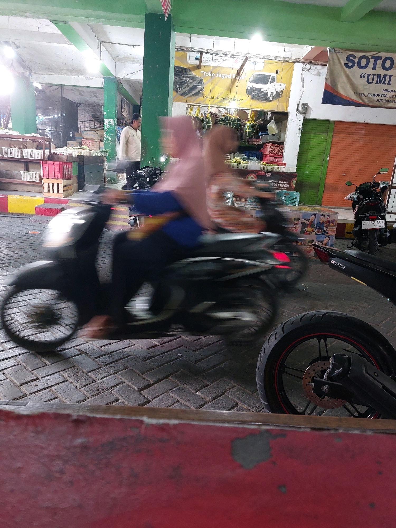 In Sumuragung, Indonesia, a market scene unfolds with a focus on motion blur. A person on a black scooter is captured mid-ride, their form indistinct due to movement. The scooter itself is dark, with a visible front wheel and headlight. In the background, a man in a light-colored jacket stands near stalls displaying various goods, including white plastic baskets and red crates. Banners with text and images are visible on the walls, suggesting commercial activity. Another motorcycle is parked on the right side of the image, identifiable by its license plate and taillight. The ground is paved with interlocking bricks.