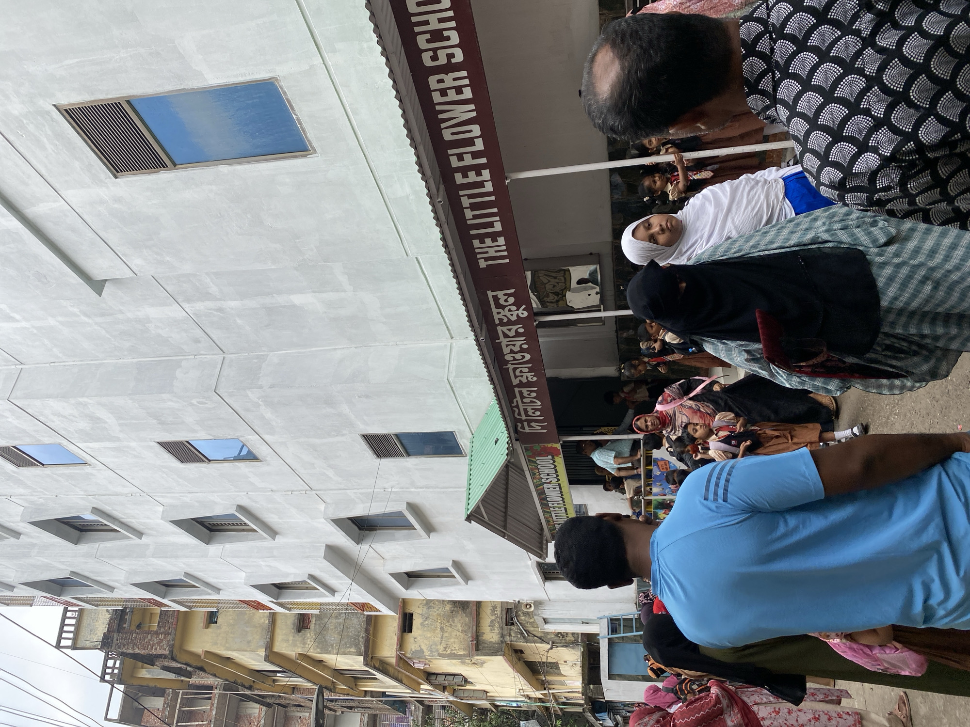 The image depicts the entrance to The Little Flower School in Dhaka, Bangladesh, with a gathering of people, mostly women and children, in front of the building. The school is a multi-story building with a light-colored facade and square windows. The school's name is displayed in English and Bengali on a sign above the entrance. People are dressed in various clothing styles, including traditional Islamic attire. The setting appears to be a street in a densely populated urban area, with other buildings visible in the background.