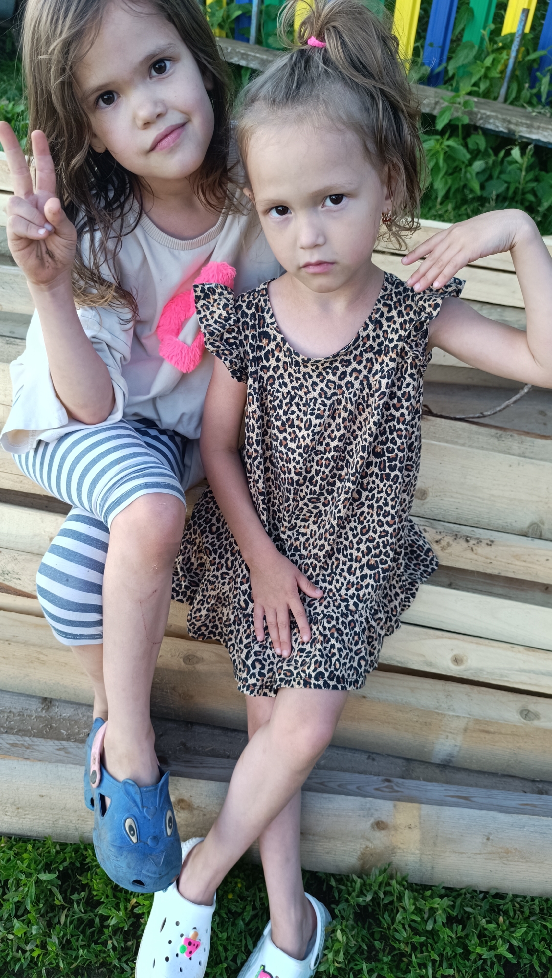Two young girls are sitting on wooden planks outdoors. The girl on the left has long, brown hair and is wearing a light-colored shirt with blue and white striped leggings. She is holding up two fingers in a peace sign. The girl on the right has her hair in a ponytail and is wearing a leopard-print dress. She has her arm resting on her shoulder. Both girls are wearing clogs on their feet. The background shows greenery and a wooden fence. The scene appears to be in a residential area, possibly a backyard or a park.
