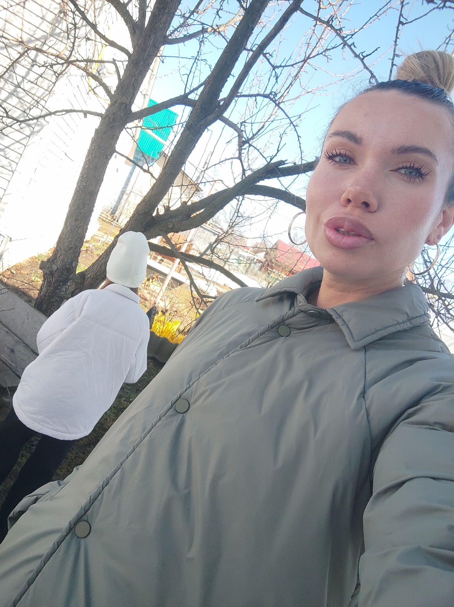 The image appears to be a selfie taken outdoors in a location possibly in Votkinsk, Russia, based on the architectural style and vegetation present in the background.
A woman with fair skin, blonde hair in a bun, and light blue eyes is in the foreground. She is wearing silver hoop earrings and a muted green, button-down puffer jacket.
Behind her to the left, partially obscured by tree branches, is another person wearing a white padded jacket and a white beanie. They are standing or sitting on a wooden structure or bench.
The backdrop includes a leafless tree with visible branches, a building with a light-colored facade, and a glimpse of what seems to be a residential area with pitched roofs. The sky is pale blue.
The lighting suggests it is daytime.