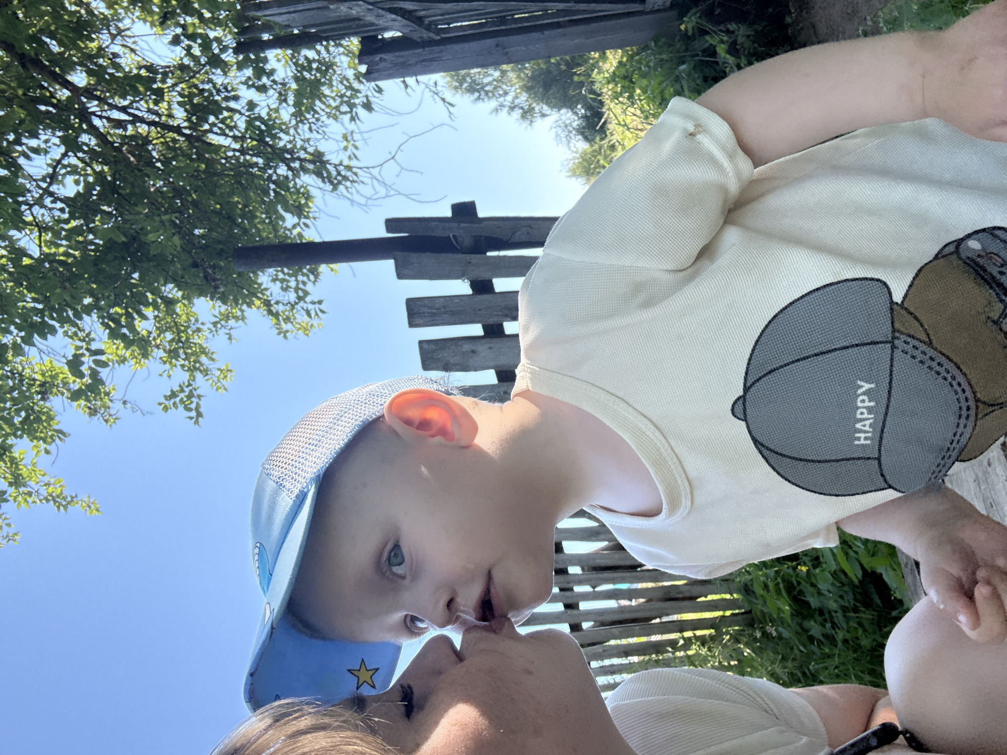 The media file is a close-up, low-angle shot of a woman and a child outdoors. The subjects are in the foreground, with their faces close to each other, in what appears to be an affectionate moment. The child, wearing a blue cap with a star and a light-colored t-shirt featuring a graphic of a cap with the word "HAPPY" visible, is looking towards the woman. The woman's face is partially visible, with her lips touching the child's.
The background suggests a rural setting, with lush green foliage and weathered wooden structures, possibly a fence or an old building. The sky is a clear, bright blue, indicating it is daytime and likely sunny. The overall mood seems warm and loving, due to the close proximity and facial expressions of the subjects. The image's orientation is rotated, with the subjects appearing upside down relative to a typical upright orientation.
Visible text includes "HAPPY" on the child's t-shirt graphic. The location context provided is Ляльшур, Russia.