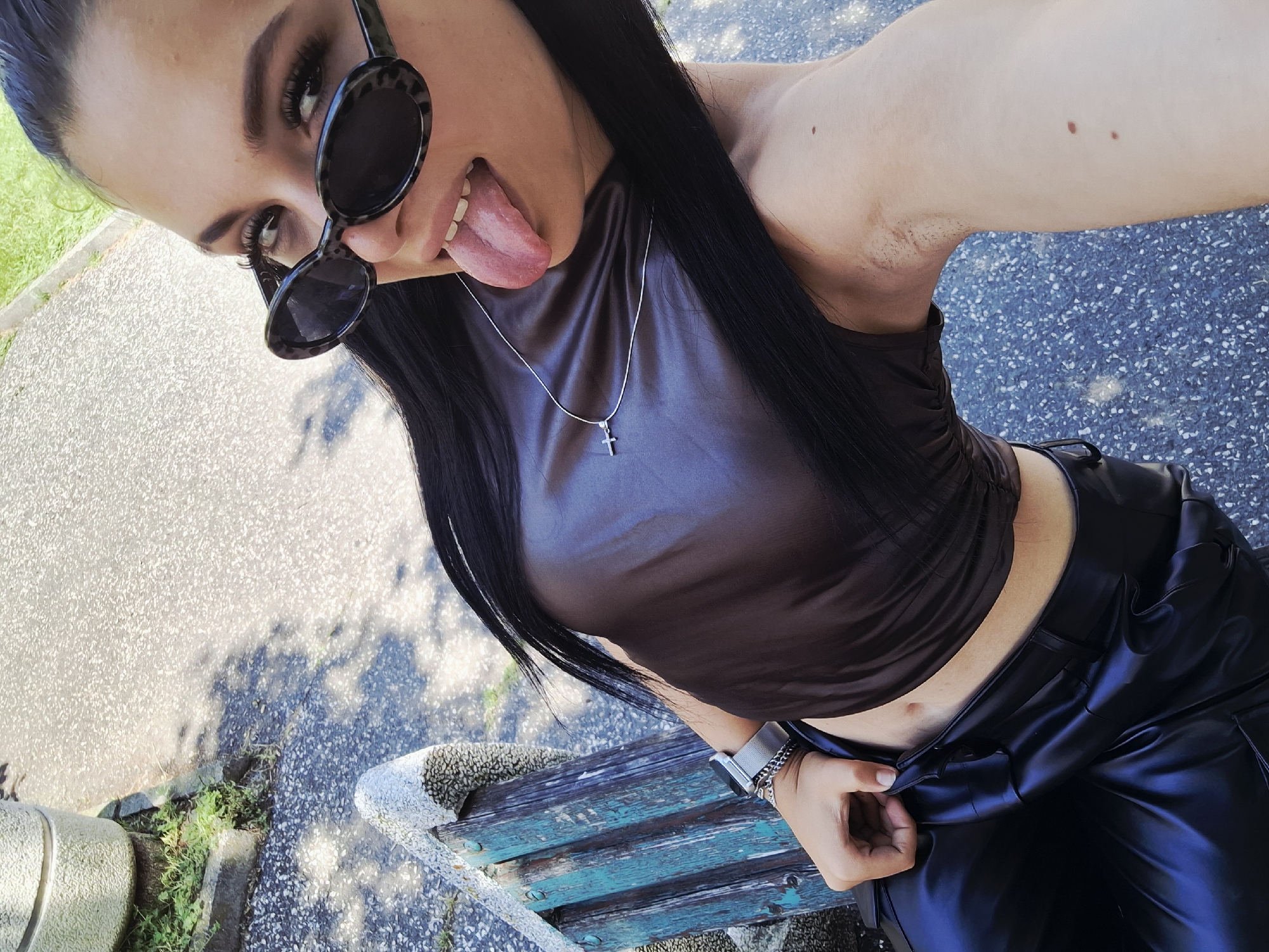 A young woman takes an overhead selfie outdoors in Mezdra, Bulgaria, under bright daylight. She has long, dark hair and wears leopard-print sunglasses, with one lens partially lowered, revealing her visible eye and dark, well-groomed eyebrows and eyelashes. She playfully sticks her tongue out towards the camera.
She is dressed in a dark brown, possibly strapless or spaghetti-strap, crop top that reveals her midriff and navel. A thin silver chain with a small silver cross pendant hangs around her neck. On her left wrist, she wears a silver watch. Her left hand is positioned near the waistband of her shiny black, leather-look pants, which appear high-waisted. Visible stubble is present in her left armpit, and a few small moles are seen on her upper arm/shoulder.
The background shows a light-colored paved surface with a speckled texture, typical of an outdoor public area like a park or street. Strong shadows cast on the ground indicate a sunny day. A distressed wooden bench with chipped blue paint is partially visible beside her, and green grass peeks out from the edge of the paved surface. The overall impression is casual and playful. There is no visible text in the image.