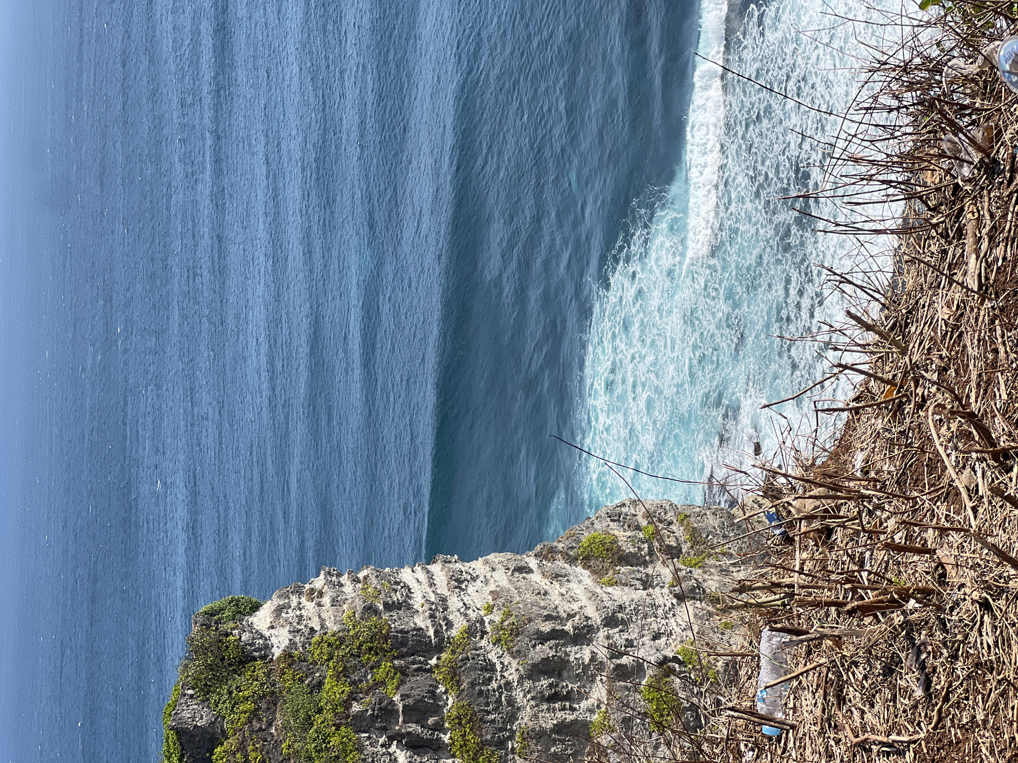The image depicts a rugged cliffside overlooking the ocean in Kuta Selatan, Indonesia. The cliff is composed of stratified rock with patches of green vegetation clinging to its surface. Below, the deep blue ocean is marked by white-capped waves crashing against the shore, creating a foamy turquoise and white pattern. The foreground of the image is dominated by dry, brown brush and debris, including discarded plastic bottles, which suggests a less-than-pristine environment at the cliff's edge. The sky is a clear, bright blue, indicating a sunny day.
