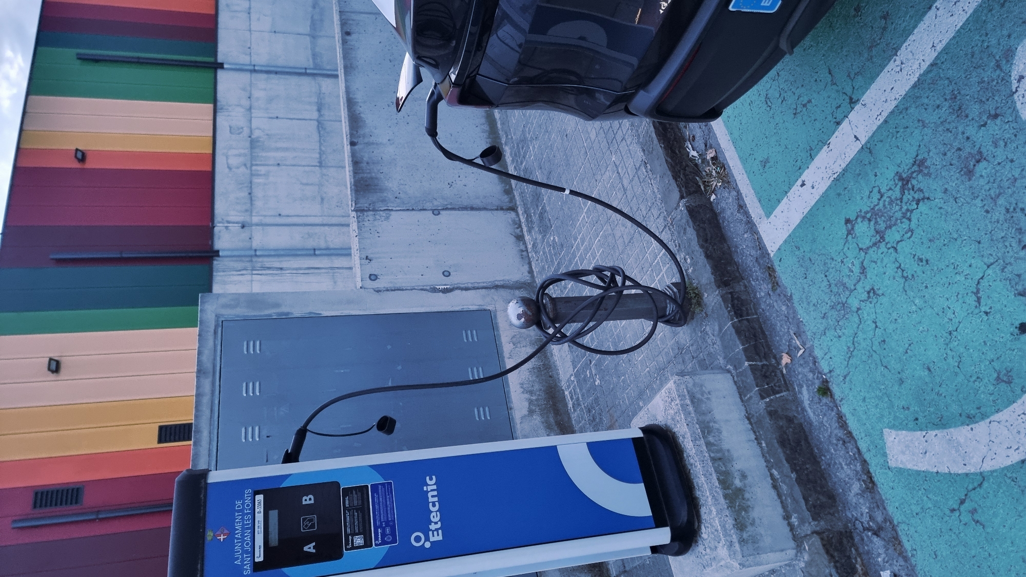 An electric vehicle charging station is in operation in the city of Sant Joan Les Fonts, Spain. A black electric car is parked in a designated charging spot, indicated by a teal-colored painted area on the ground with white markings. A black charging cable extends from the charging station, which is a blue and white pedestal unit with the Etecnic logo and "AJUNTAMENT DE SANT JOAN LES FONTS" text on its side. The cable is plugged into the car. The charging cable is coiled around a short, dark metal bollard located between the charging station and the car. In the background, a concrete wall runs along the property, and behind that, a building facade is brightly colored with vertical stripes of red, orange, yellow, green, and blue. The ground around the charging station is paved with square tiles. The sky is overcast.
