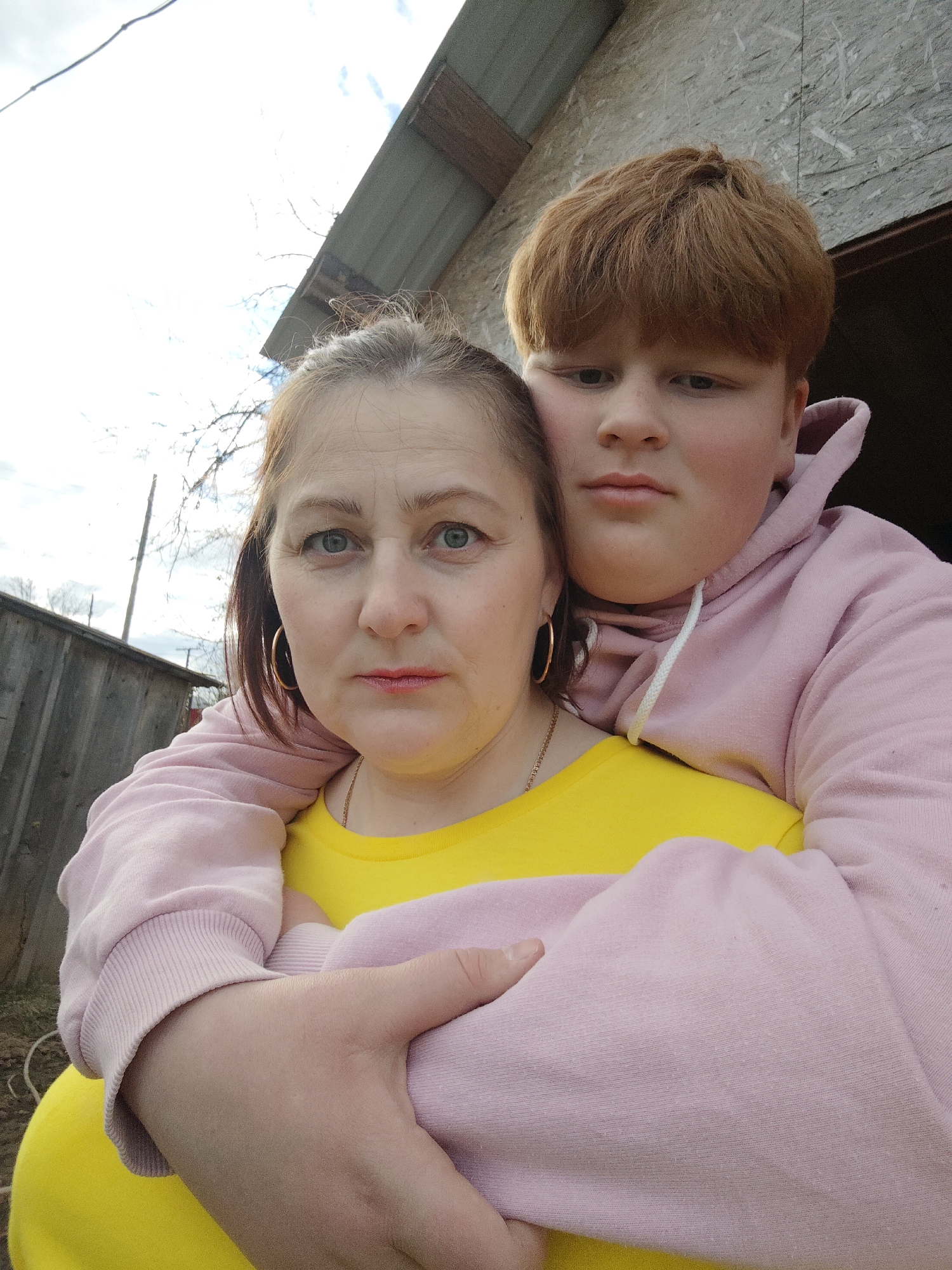 A close-up outdoor portrait captures a woman in the foreground, embraced from behind by a boy. Both subjects look directly at the camera with neutral, slightly serious expressions. The woman, in the foreground, has light-colored eyes, and her hair, possibly pulled back, shows lighter streaks. She wears a bright yellow t-shirt, gold hoop earrings, and a thin gold chain necklace, with red lipstick adorning her lips. Behind her, the boy, with reddish-brown hair cut in a bowl style, rests his head on her right shoulder and wears a light pink hooded sweatshirt. His arms are wrapped around the woman's torso.
The setting is outdoors, under a bright, partly cloudy sky, indicating daytime. In the background, to the left, a weathered wooden fence is visible. To the right and behind the subjects, parts of a simple structure are seen, featuring what appears to be a corrugated metal roof and wooden or OSB siding. The scene suggests a rustic or residential outdoor environment, consistent with the provided location context of Фоки, Russia. There is no visible text in the image.