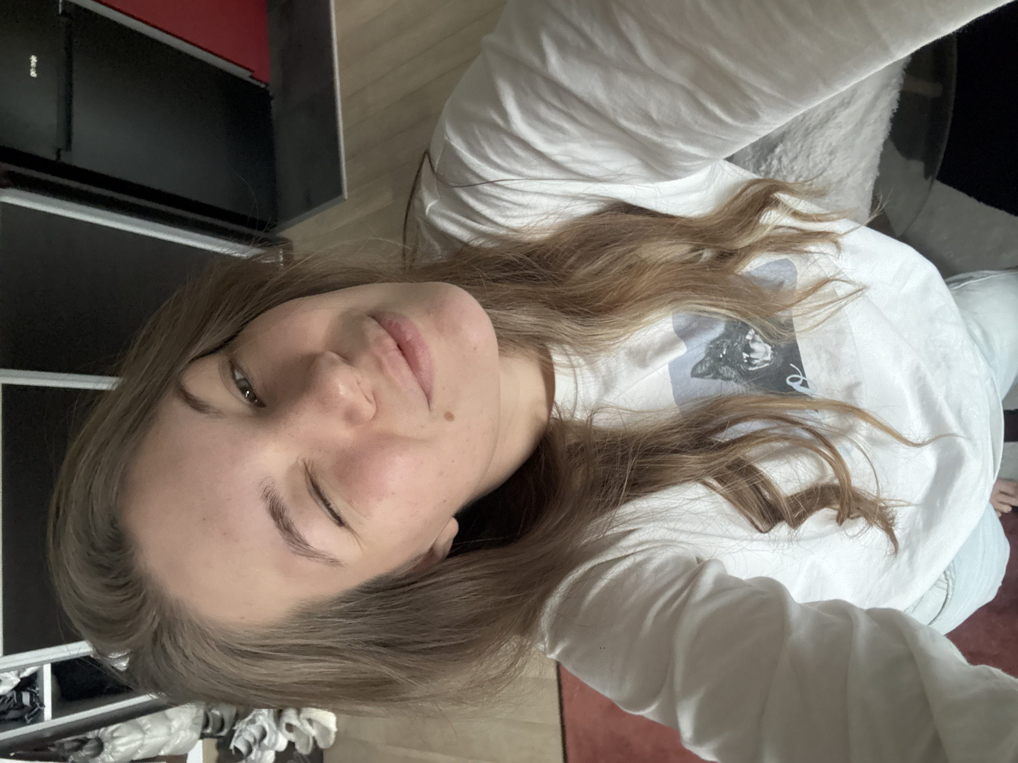 A woman with fair skin and brown hair, wearing a white long-sleeved shirt, is lying down. A grey cabinet is behind her to the left. The floor is visible. The location is not discernible from the image.
