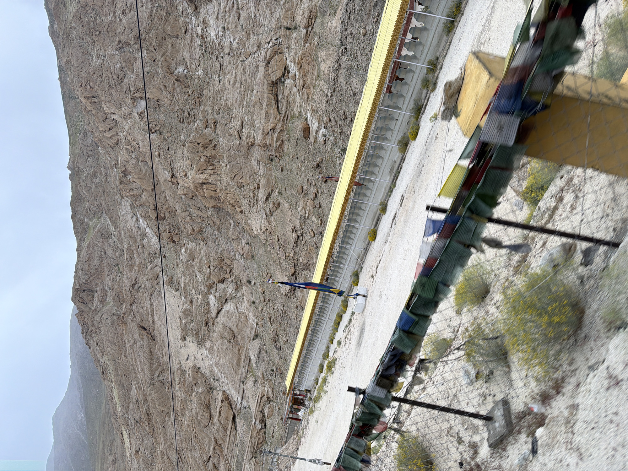 This image depicts a mountainous landscape with a long, low-lying structure featuring a yellow roof supported by white pillars. In front of this structure, there is a sandy area with sparse vegetation and a fence adorned with colorful prayer flags. A flagpole with a flag is also visible near the structure. The background is dominated by a steep, rocky mountain slope. The scene appears to be located in Kungyamdo, India, a city known for its mountainous terrain and cultural heritage. The overall impression is one of a serene, spiritual setting amidst a rugged natural environment.