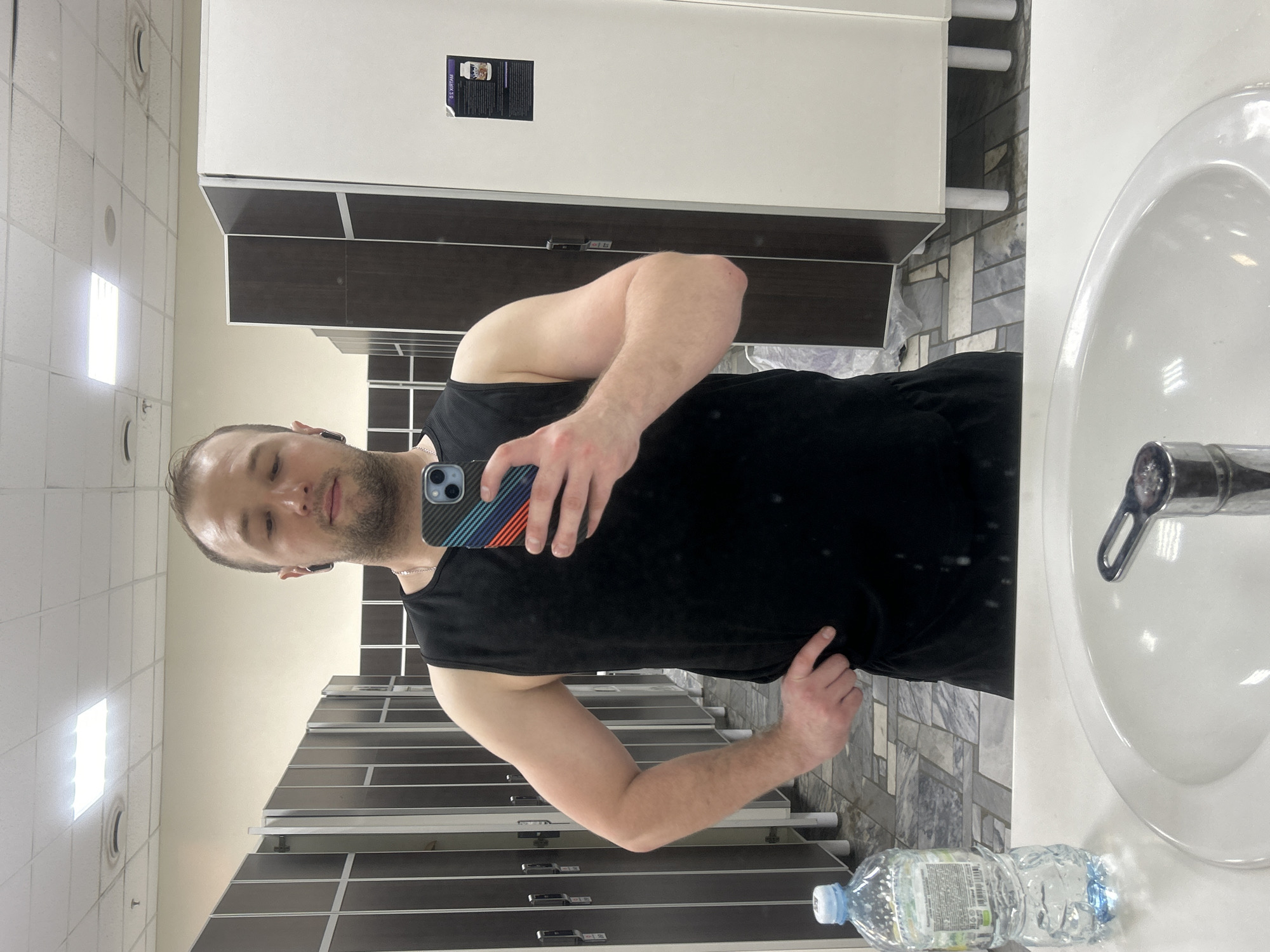 A man is taking a selfie in a bathroom. The location is likely in Chaikovsky, Russia, based on the content of a small sign visible on a locker. The man is wearing a black tank top and holding a phone with a colorful case. He has short hair and a beard. A sink and faucet are visible to his right. There are lockers along the wall behind him, and two plastic bottles are on the counter to the right. The floor appears to be tiled.