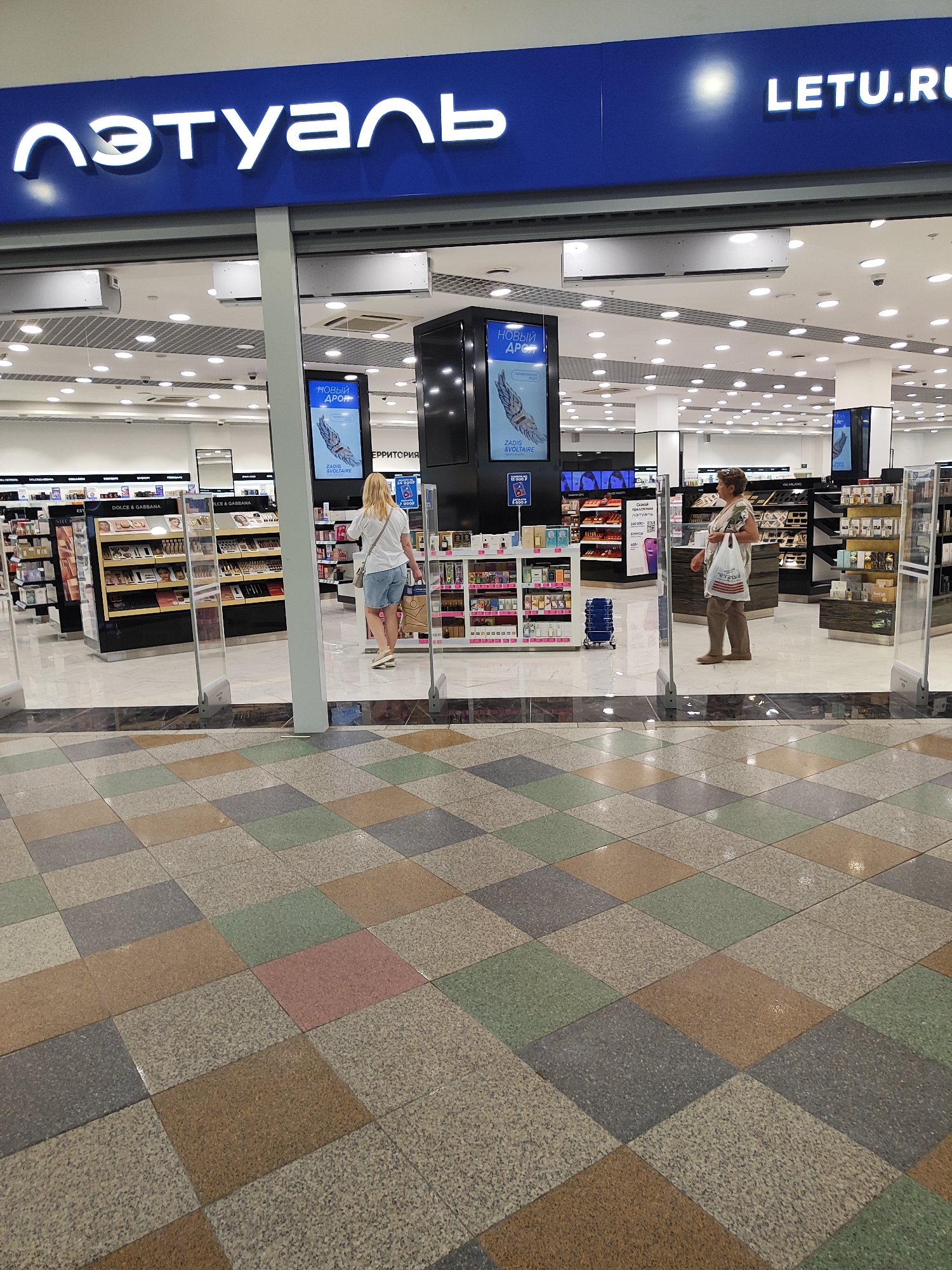 A wide shot captures the entrance of a L'Etoile beauty store. The store's blue and white sign is prominent, displaying "l'etu але" and "LETU.RU". Inside, shelves are stocked with cosmetics and perfumes. Two women are visible: one, wearing a white t-shirt and denim shorts, browses the merchandise. The second woman, in a patterned shirt and brown pants, carries a shopping bag and stands near a display. The store's interior is brightly lit, with numerous ceiling lights. The floor in front of the store is tiled in a diamond pattern of various earth tones, including beige, brown, gray, and green. The scene is indoors, likely in a shopping mall.