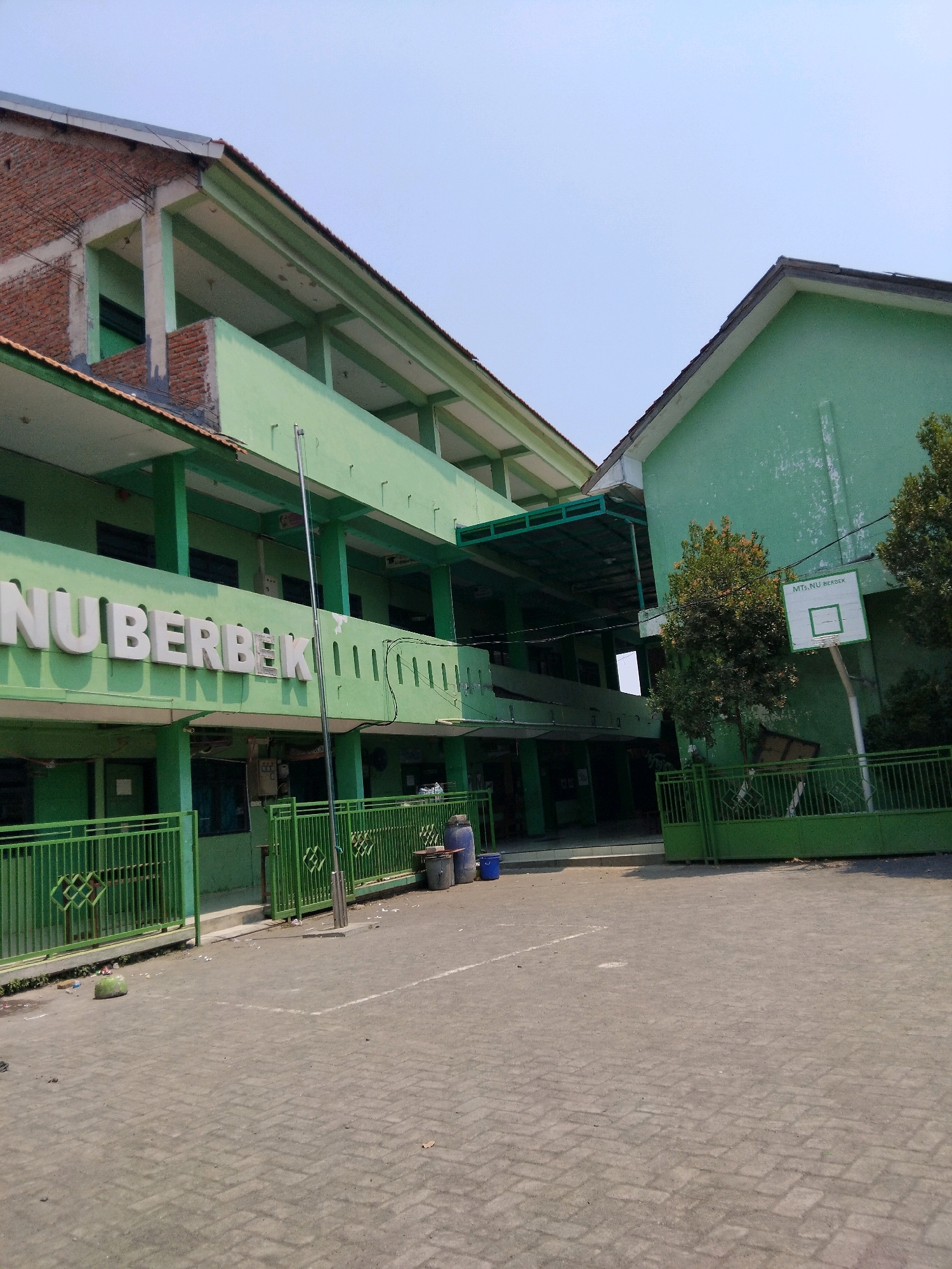 In the city of Kepuh Kiriman, Indonesia, a multi-story building, likely a school, stands with its exterior painted a vibrant green. The building features multiple levels with open walkways and balconies, suggesting an outdoor corridor design. Large, white, three-dimensional letters spelling "NU BERBEK" are prominently displayed on the facade of one section of the building. A basketball hoop with a backboard is mounted on a wall of another section of the building, with the words "MTS. NU BERBEK" visible on the backboard. The ground in front of the building is paved with interlocking gray bricks. A green metal fence runs along the perimeter of the building, bordering the paved area. Several large blue and gray plastic barrels are situated near the fence. A flagpole stands tall in the paved area. The sky above is clear and blue, indicating a sunny day. The overall scene depicts an educational facility in an urban or semi-urban environment.