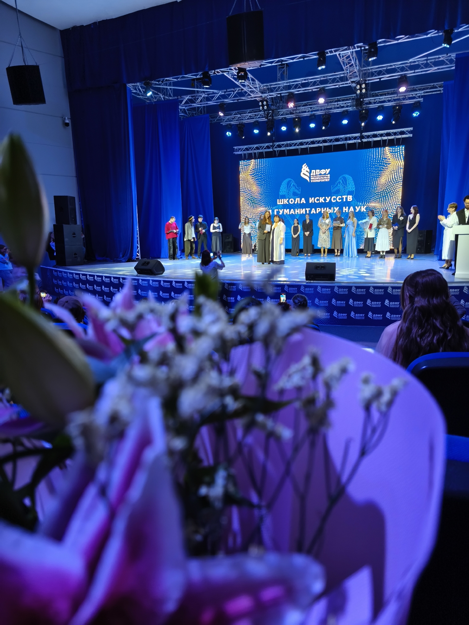 This image depicts an indoor awards ceremony or graduation at the Far Eastern Federal University (FEFU) in Russky Island, Primorsky Krai, Russia. A group of individuals, likely students and faculty, are on stage receiving recognition or presenting awards. They are dressed in formal attire, including some in academic regalia. The stage is adorned with blue curtains and a large LED screen displaying the university's logo and the text "ШКОЛА ИСКУССТВ ГУМАНИТАРНЫХ НАУК" which translates to "School of Arts and Humanities." The audience is seated in a dimly lit hall, with some individuals in the foreground blurred and out of focus, suggesting the photo was taken from within the audience. The overall atmosphere appears celebratory.