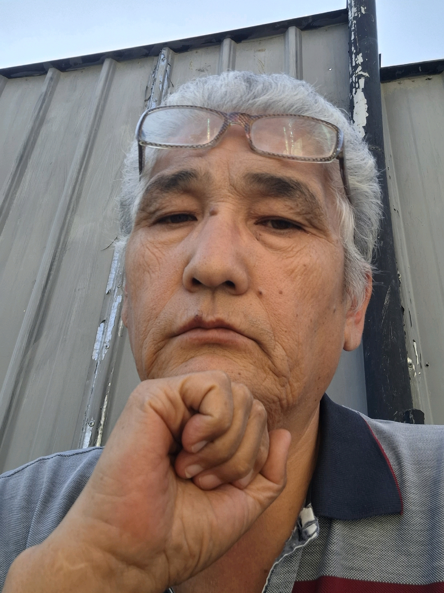 A close-up, low-angle, outdoor selfie features an elderly East Asian male with graying hair and glasses, resting his chin on his hand. He is wearing a grey, red, and navy collared shirt. The background consists of a corrugated metal wall and a sliver of bright sky. There is no identifiable information within the image to suggest a specific location in Almaty, Kazakhstan.