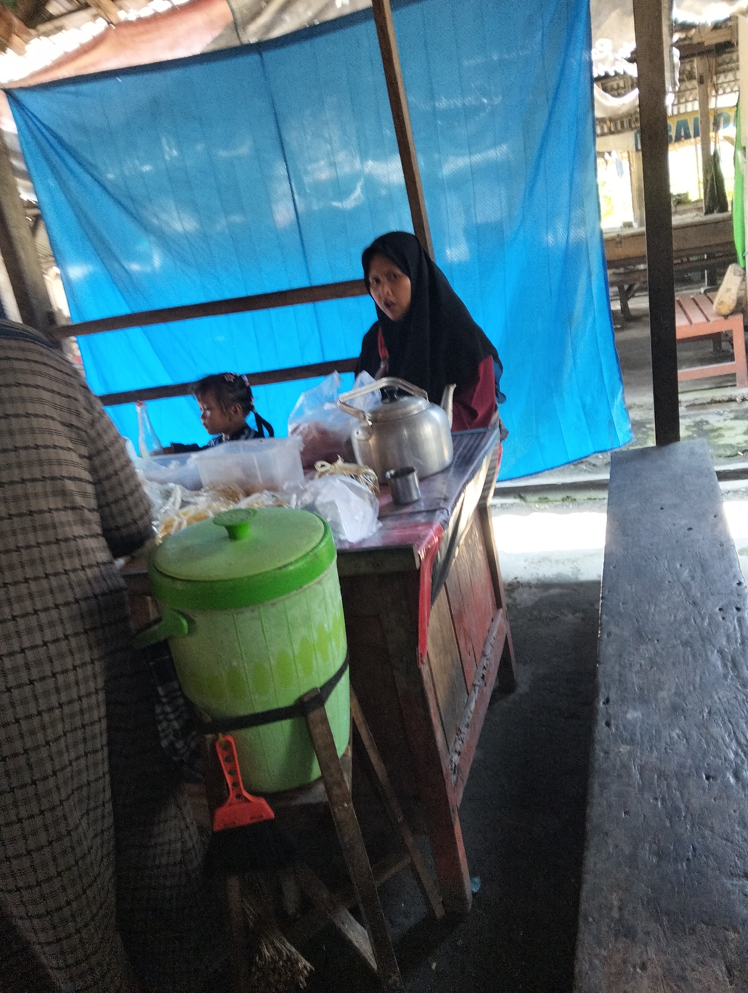 A woman wearing a black hijab and a red top stands behind a wooden counter at a street-side stall in Muncar, Indonesia. She is looking towards the camera. To her left, a young girl with braided hair is visible. The stall is covered by a blue tarp. On the counter, there is a silver teapot, a small metal cup, plastic bags with food items, and a green bucket with a black strap and a small orange broom. A checkered patterned garment is draped over a structure to the left of the counter, partially obscuring the scene. A wooden bench or table is positioned in the foreground. The ground appears to be concrete or a similar hard surface.